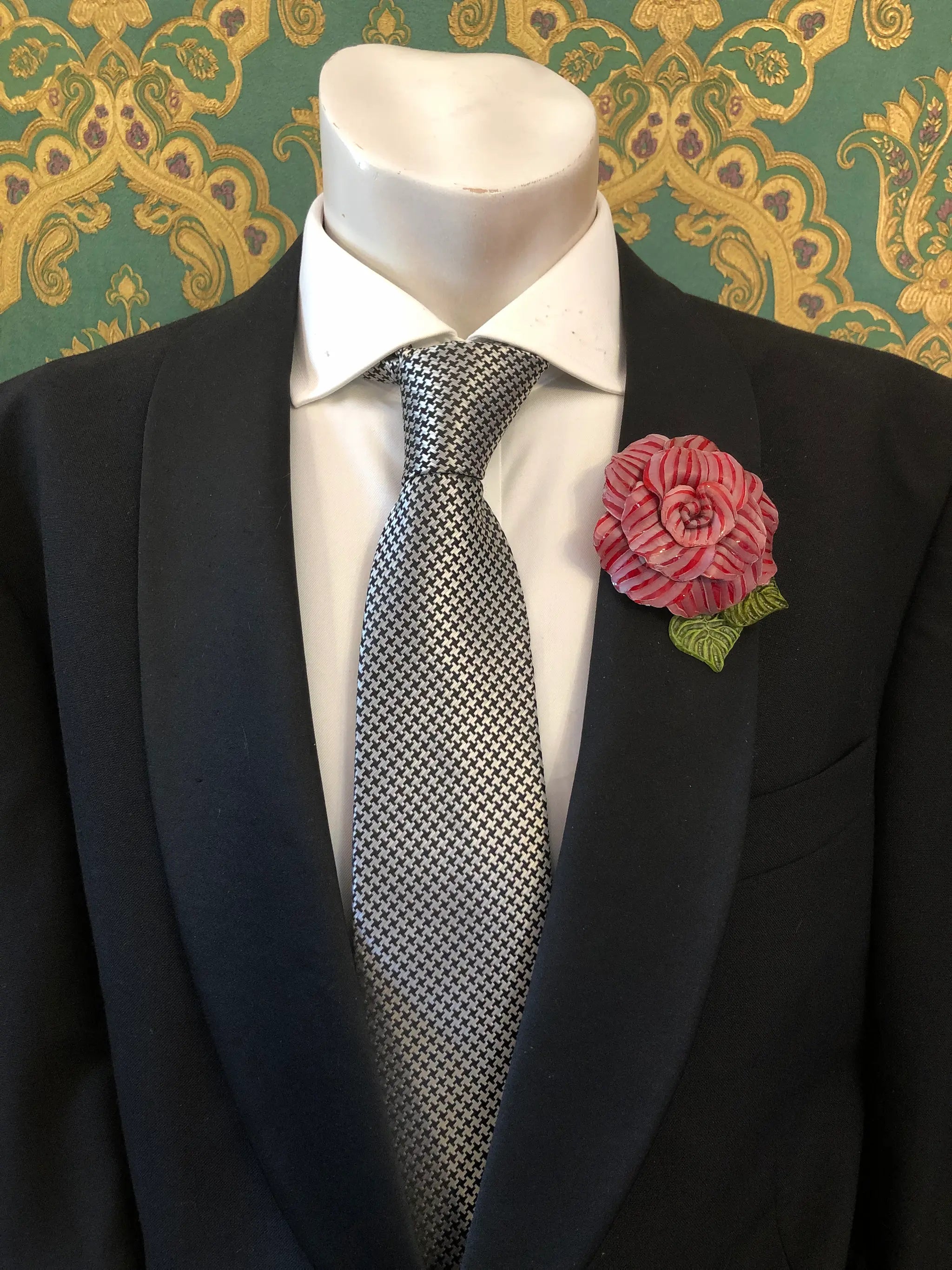 A men's formal outfit displayed on a white mannequin torso, featuring a black suit jacket, a white dress shirt, a grey and black checkered tie. Pinned on the lapel is a Broochella Men's Fabrice Camelia brooch in red with green leaves. The background is a green and gold patterned wallpaper.