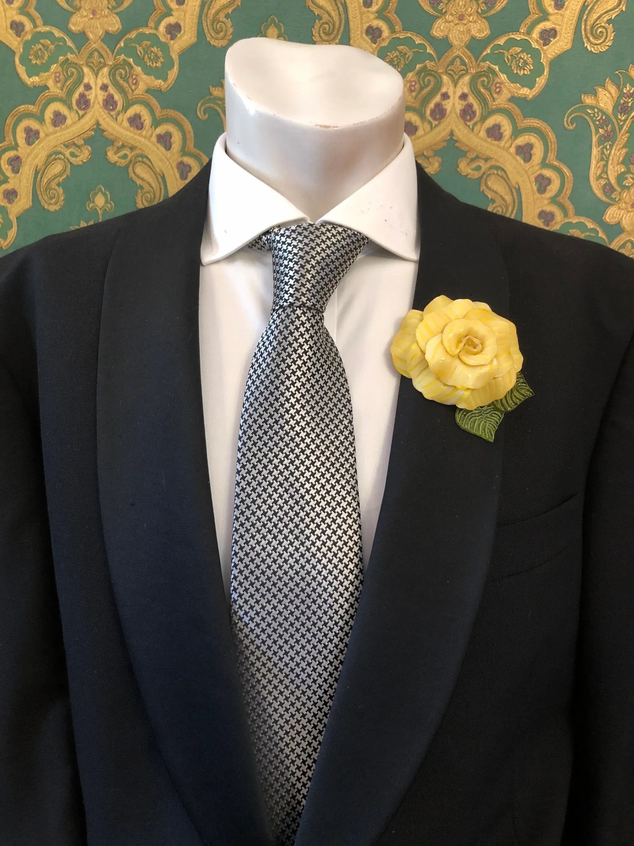 A men's formal outfit displayed on a white mannequin torso, featuring a black suit jacket, a white dress shirt, a grey and black checkered tie. Pinned on the lapel is a Broochella Men's Fabrice Camelia brooch in yellow with green leaves. The background is a green and gold patterned wallpaper.