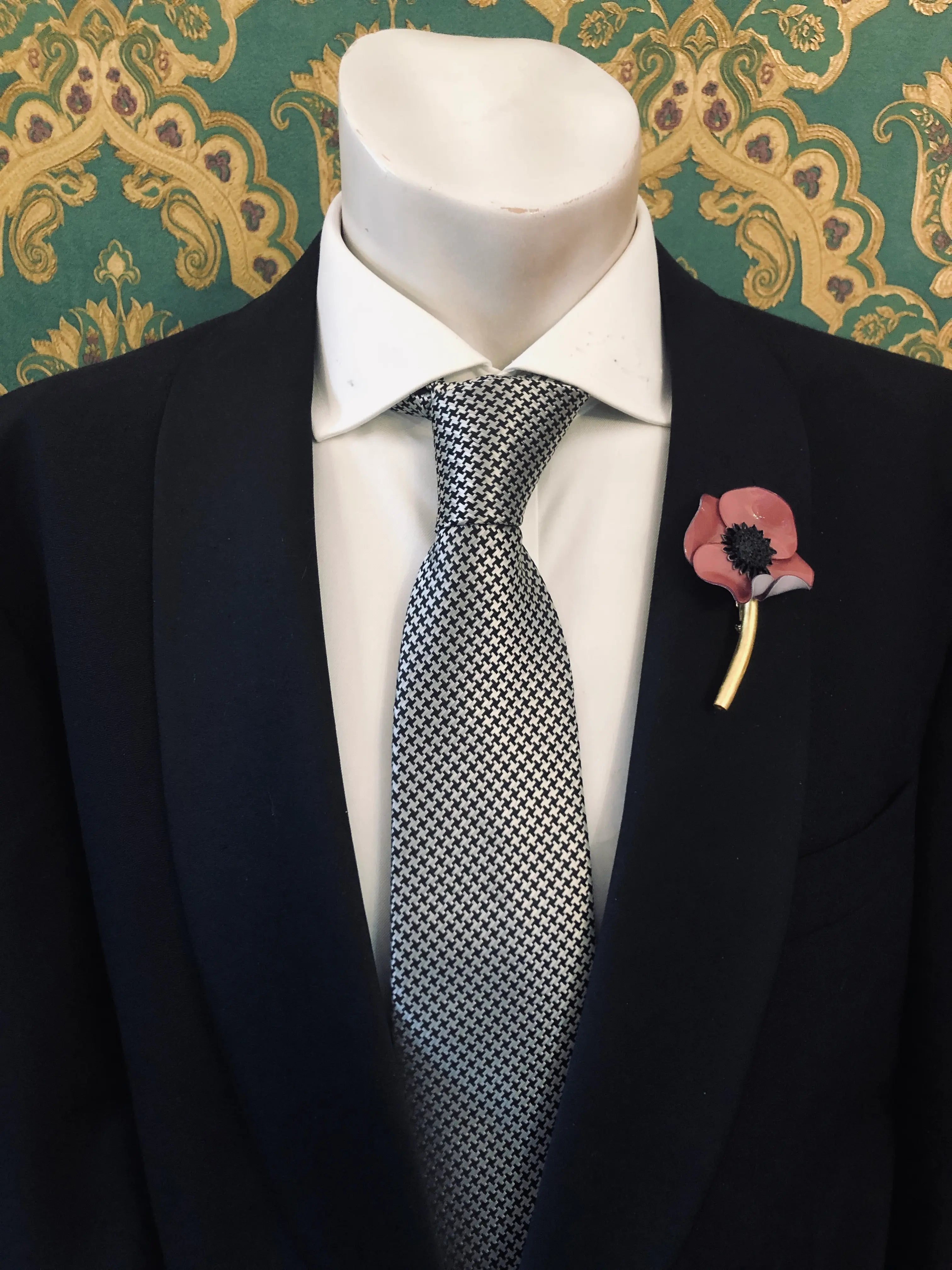 A men's necktie with a small-scale black and white geometric pattern, styled on a white dress shirt and a black suit jacket. A Broochella Men's Fabrice Paris mauve poppy lapel pin is pinned on the left lapel.
