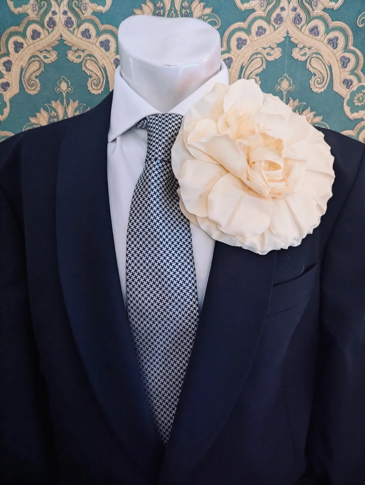 A formal outfit displayed on a white mannequin torso, featuring a black suit jacket, a white dress shirt, a grey and black patterned tie, and a large Broochella handmade men's femmina pale yellow fabric flower brooch on the left lapel. The background is a teal and beige damask patterned wall.