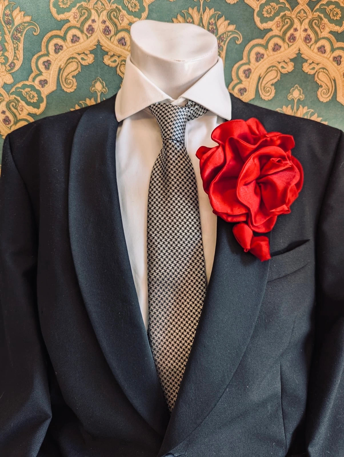 Broochella men's rosetta handmade, designer, satin flower brooch in red. Pinned on a men's mannequin wearing a black suit and a houndstooth tie. The background is green and gold damask wallpaper. 