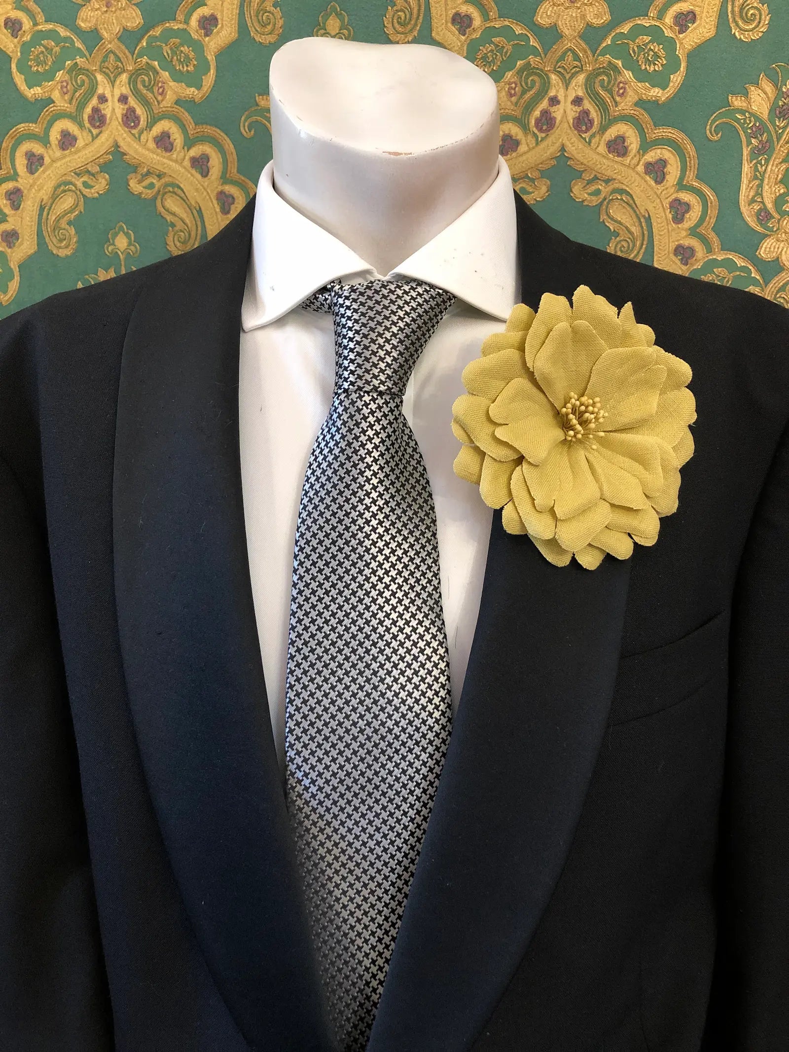 Broochella Men's Garofano handmade linen flower brooch in beige. It is pinned to a mannequin wearing a black suit, white shirt and a silver and black houndstooth tie. The background is green and gold damask wallpaper. 