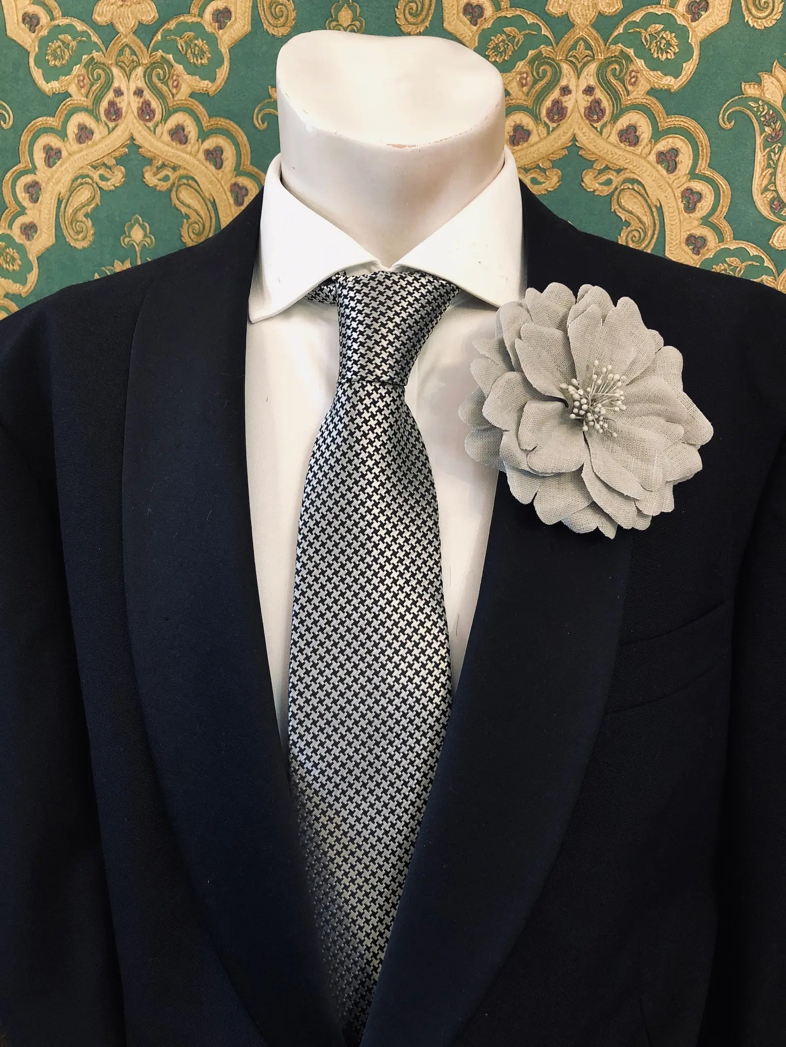 Broochella Men's Garofano handmade linen flower brooch in grey. It is pinned to a mannequin wearing a black suit, white shirt and a silver and black houndstooth tie. The background is green and gold damask wallpaper. 