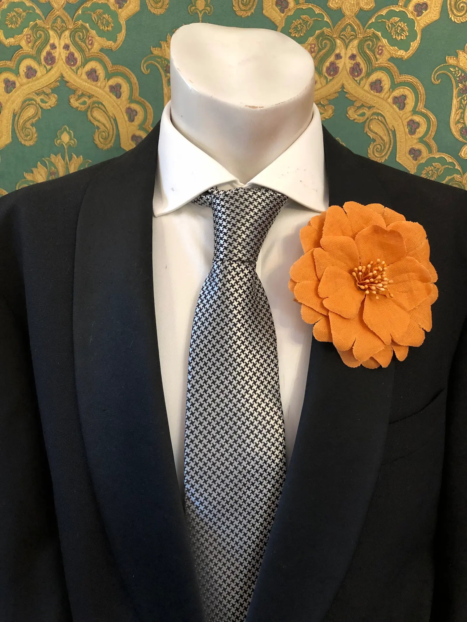 Broochella Men's Garofano handmade linen flower brooch in orange. It is pinned to a mannequin wearing a black suit, white shirt and a silver and black houndstooth tie. The background is green and gold damask wallpaper. 