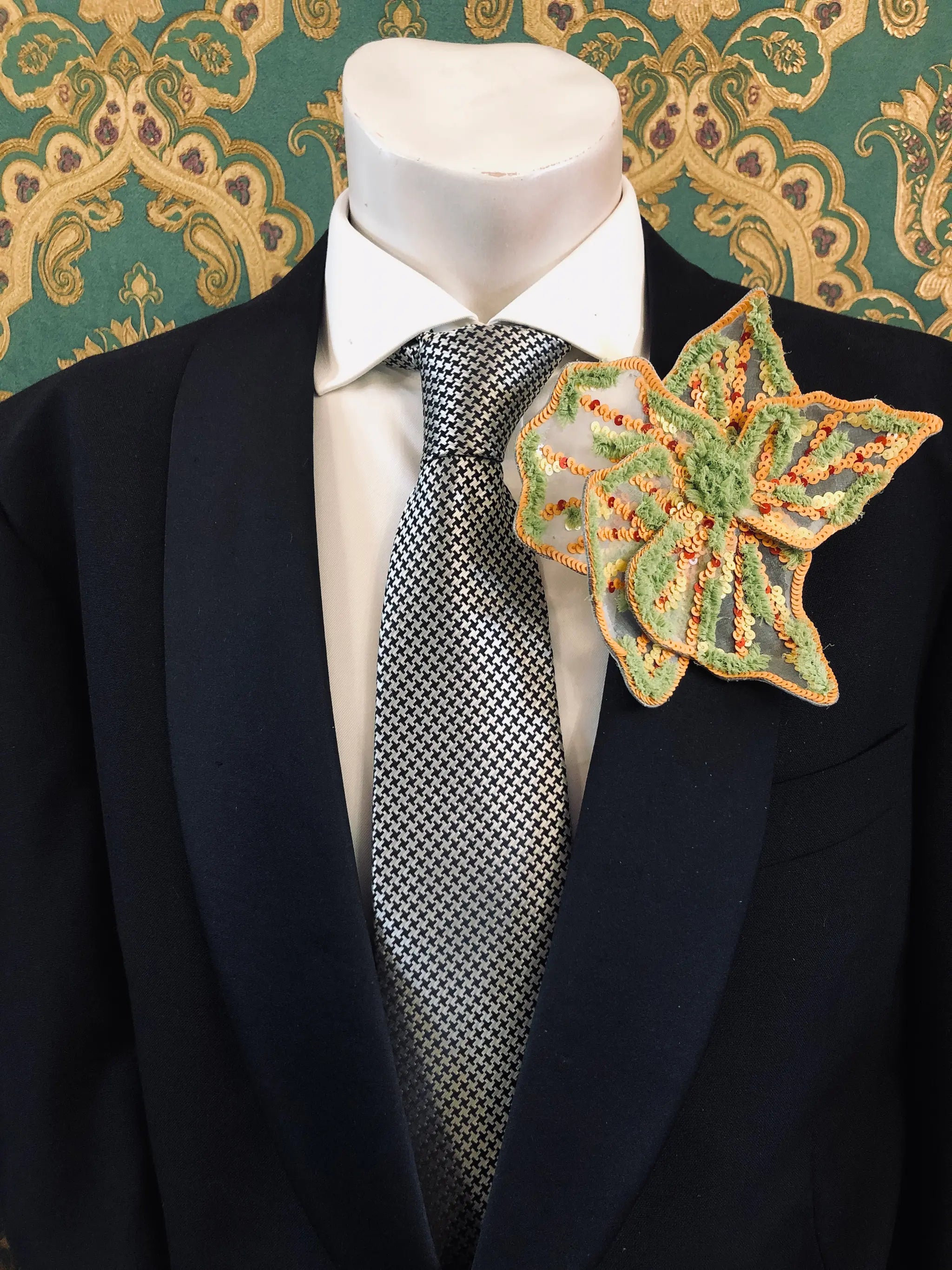 A Broochella Men's Giglio di Tigre orange and green handmade sequinned  flower brooch pin. It is styled on a black suit jacket, white dress shirt, and patterned grey tie, set against a green and gold damask wallpaper background.