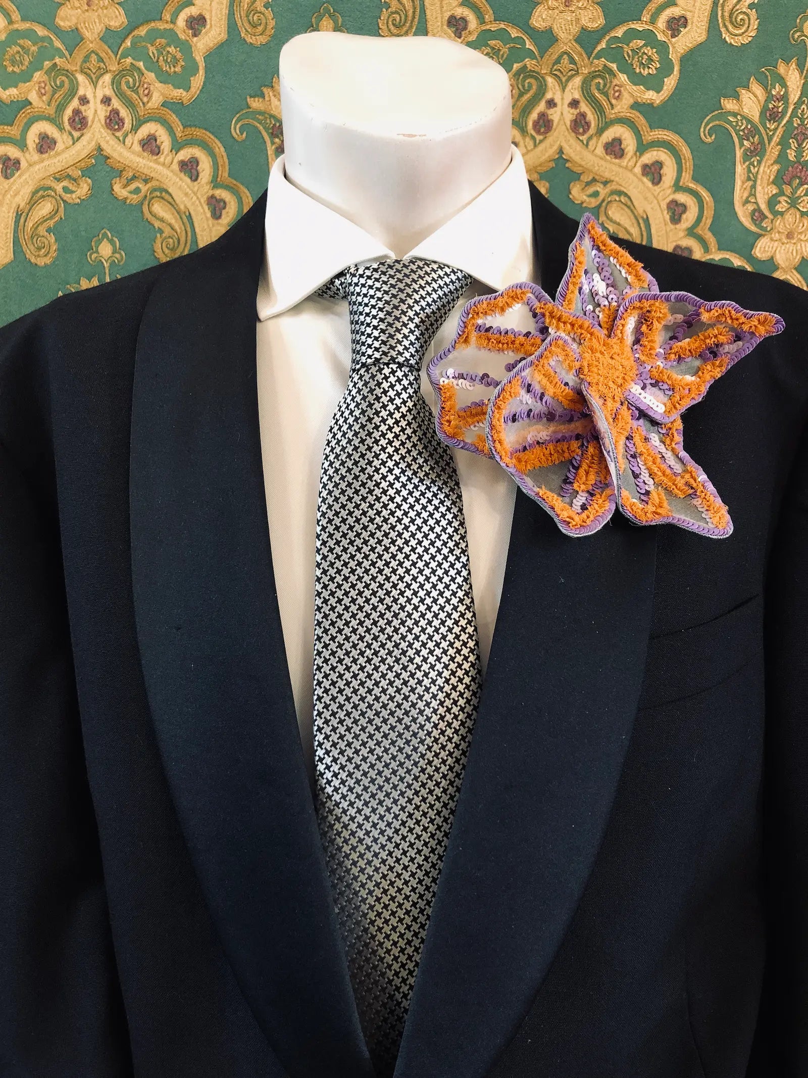 A Broochella Men's Giglio di Tigre orange and purple handmade sequinned  flower brooch pin. It is styled on a black suit jacket, white dress shirt, and patterned grey tie, set against a green and gold damask wallpaper background.