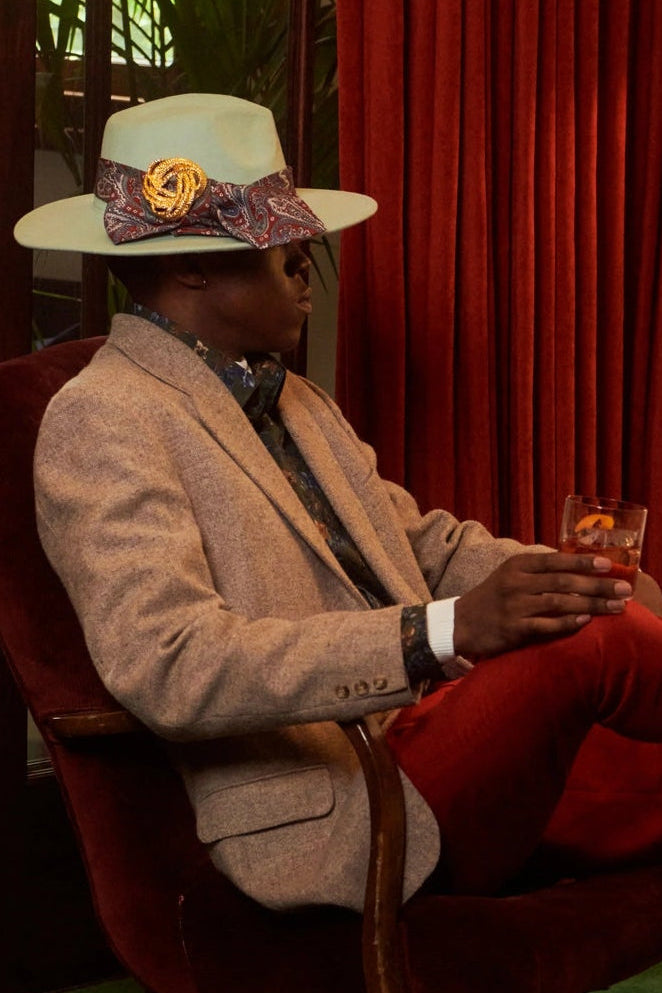 A man sits in a red armchair wearing a beige blazer, bright red trousers, a patterned white hat, and brown shoes, holding a drink in a styled interior with red curtains, green carpet, and a floor lamp. He is wearing the Broochella men's golden knot brooch on his hat