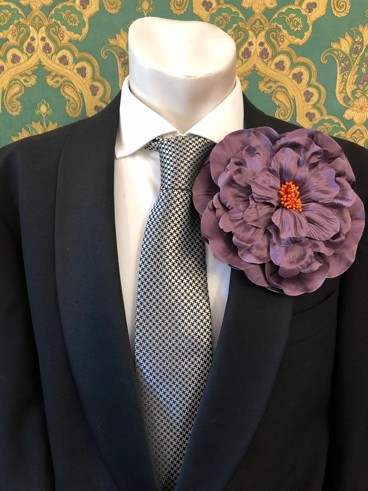 A large Broochella Men's Ibisco purple fabric flower brooch with an orange centre,  pinned to the lapel of a black suit jacket over a white dress shirt and a black-and-white patterned tie.