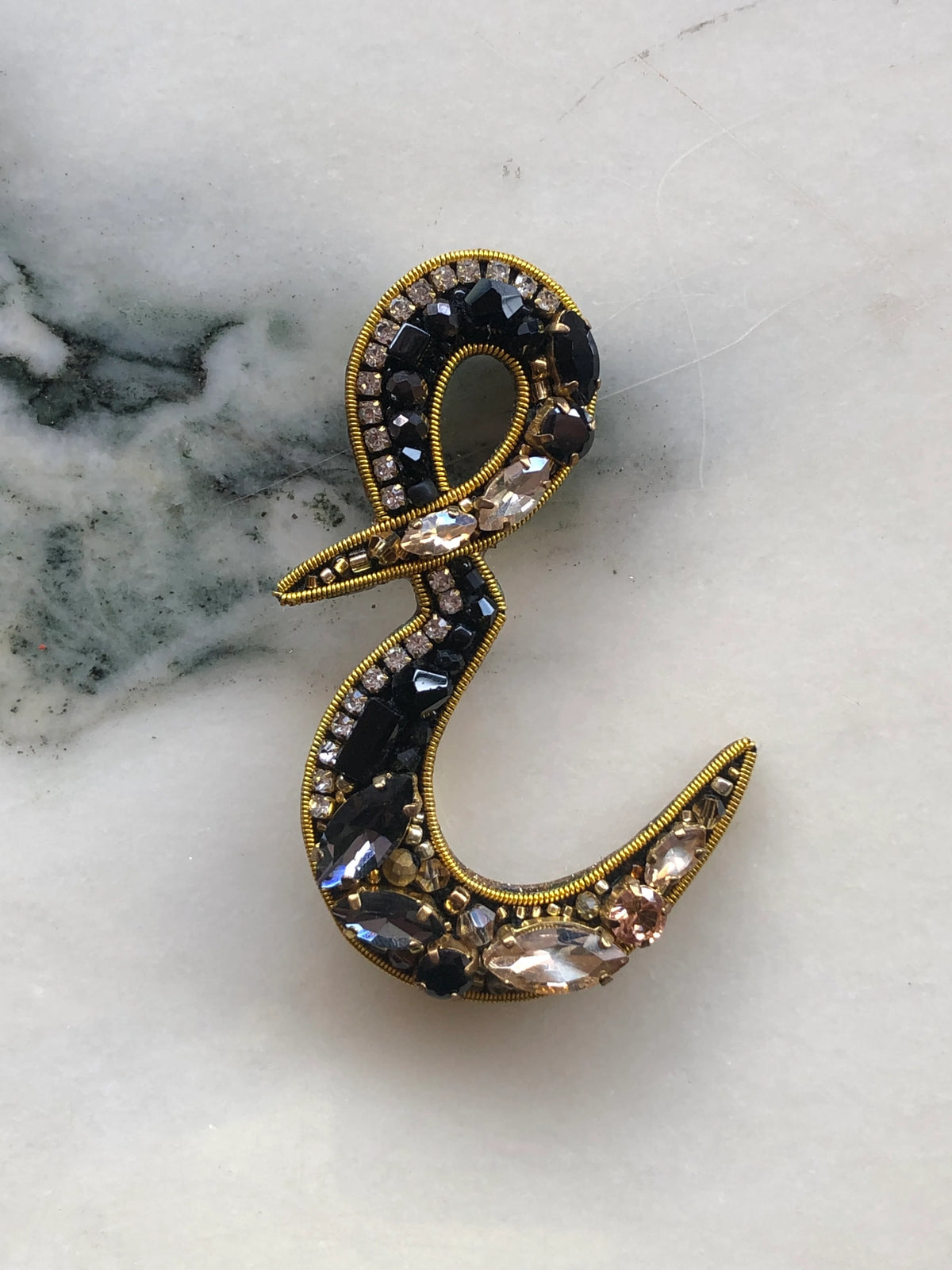 Broochella Men's Initial E Brooch embellished with black beads, clear crystals and champagne stones with gold detailing, photographed on a white marble background with soft grey veining.