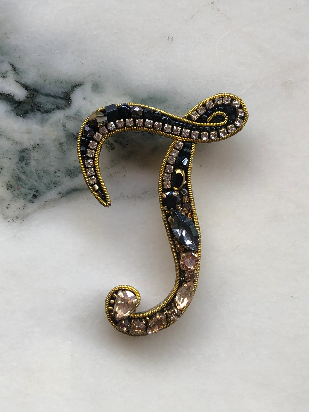 Broochella Men's Initial J Brooch embellished with black beads, clear crystals and champagne stones with gold detailing, photographed on a white marble background with soft grey veining.