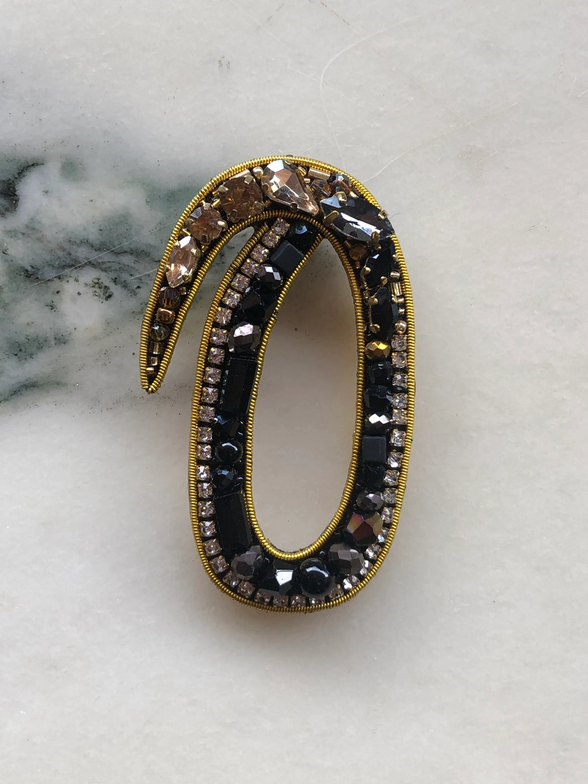 Broochella Men's Initial O Brooch embellished with black beads, clear crystals and champagne stones with gold detailing, photographed on a white marble background with soft grey veining.
