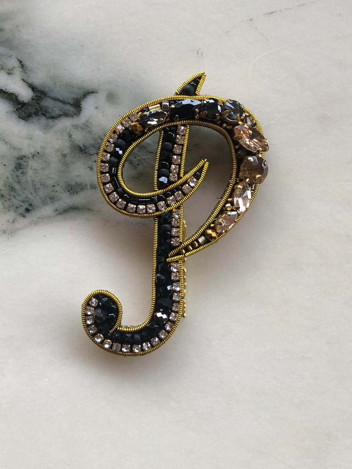 Broochella Men's Initial P Brooch embellished with black beads, clear crystals and champagne stones with gold detailing, photographed on a white marble background with soft grey veining.