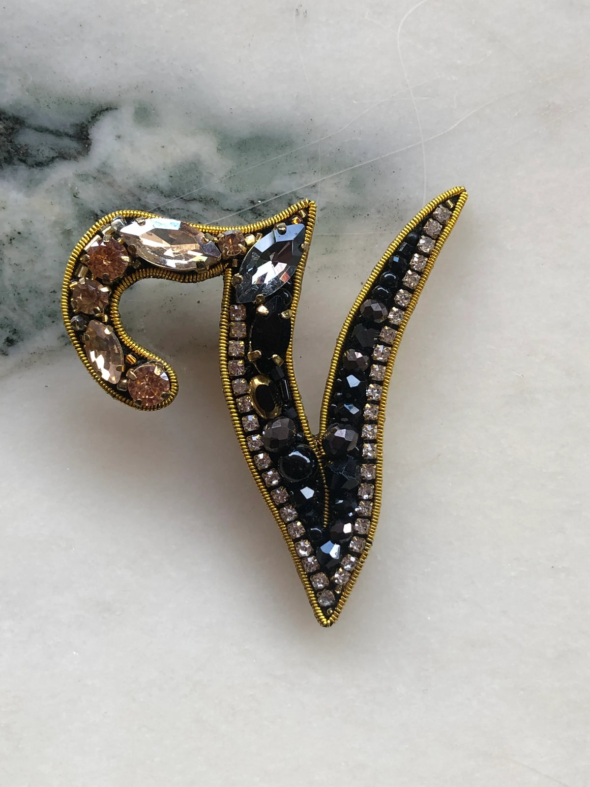 Broochella Men's Initial V Brooch embellished with black beads, clear crystals and champagne stones with gold detailing, photographed on a white marble background with soft grey veining.