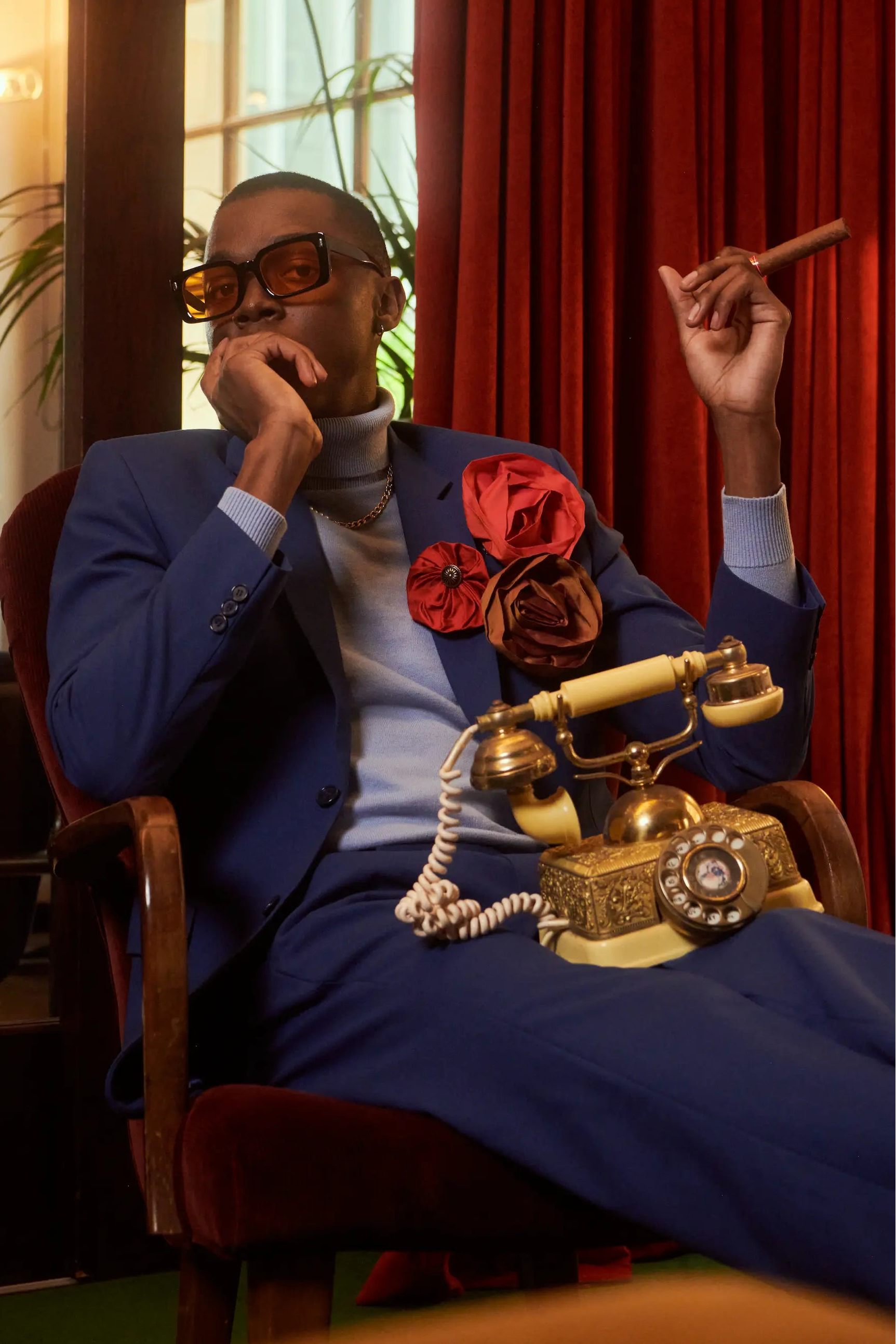 A man in a blue suit and sunglasses sits in a red armchair, holding a cigar. A vintage cream rotary telephone rests on his lap, and red curtains and warm interior lighting are visible in the background. He is wearing the Broochella Isabella Rossellini flower brooch