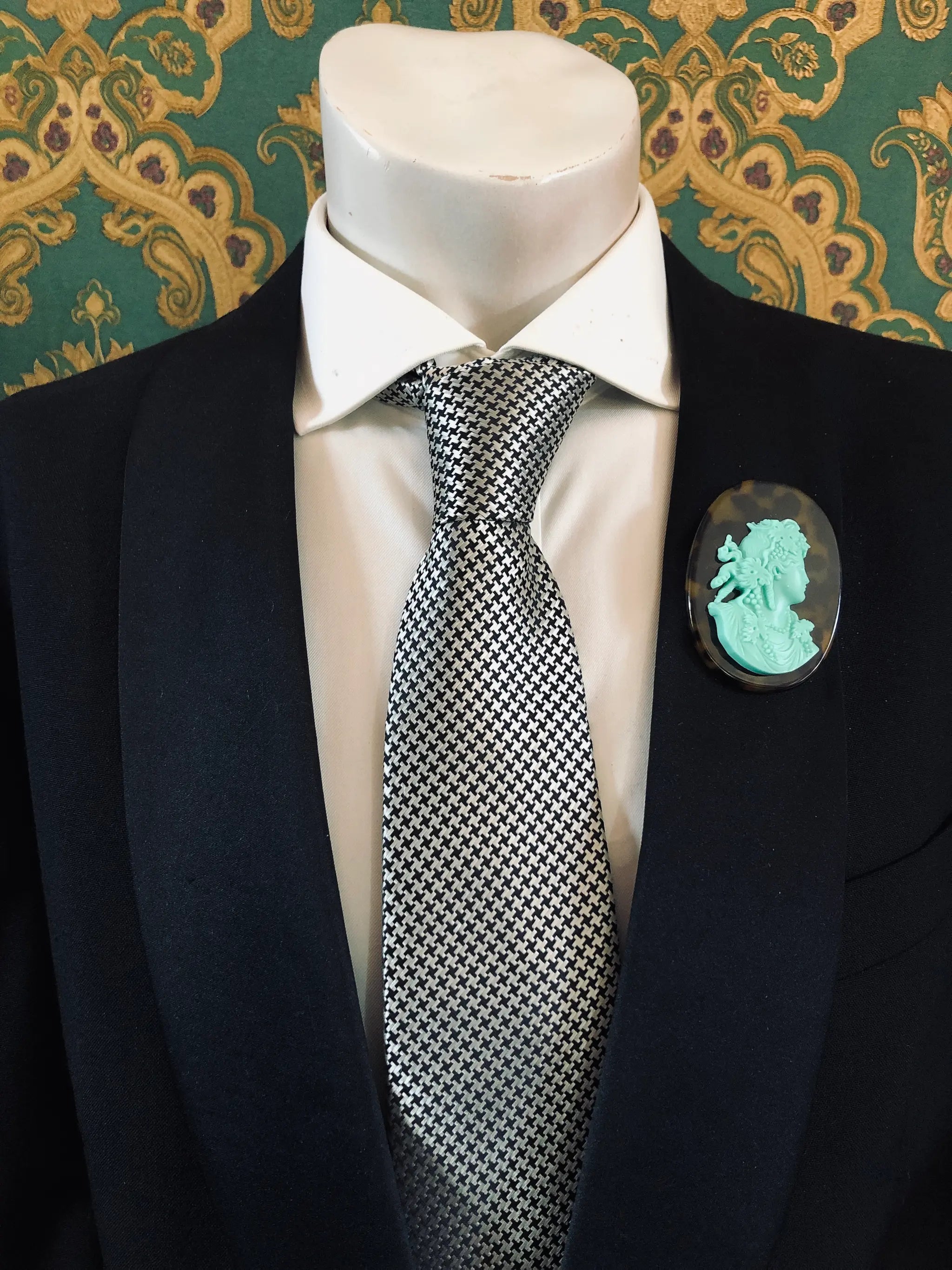 A black suit jacket on a mannequin torso, styled with a white dress shirt and a silver-and-black checkered tie. A Broochella Men's La Bella Figura bright aqua and tortoiseshell cameo oval brooch is pinned to the left chest.