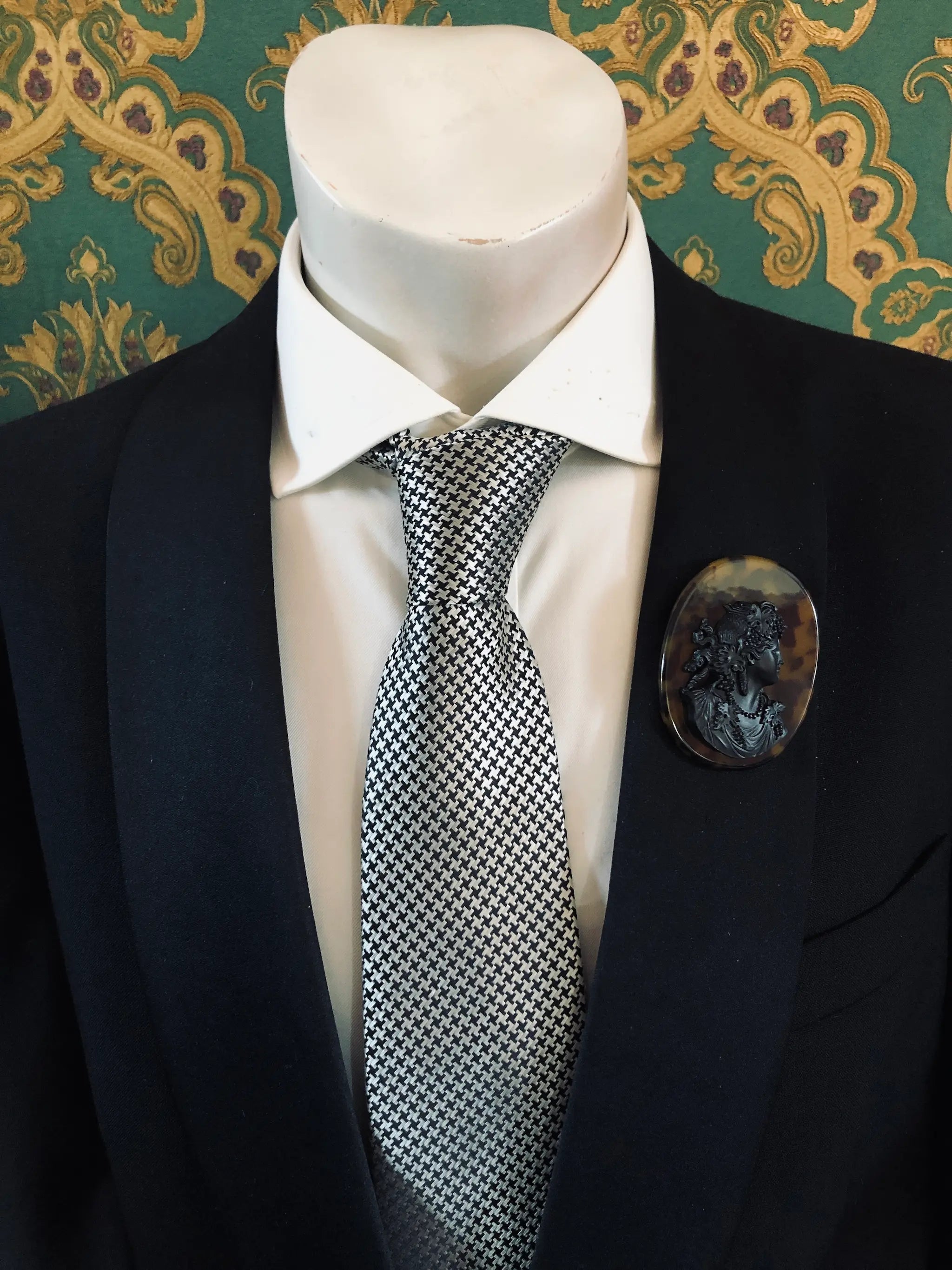 A black suit jacket on a mannequin torso, styled with a white dress shirt and a silver-and-black checkered tie. A Broochella Men's La Bella Figura black and tortoiseshell cameo oval brooch is pinned to the left chest.