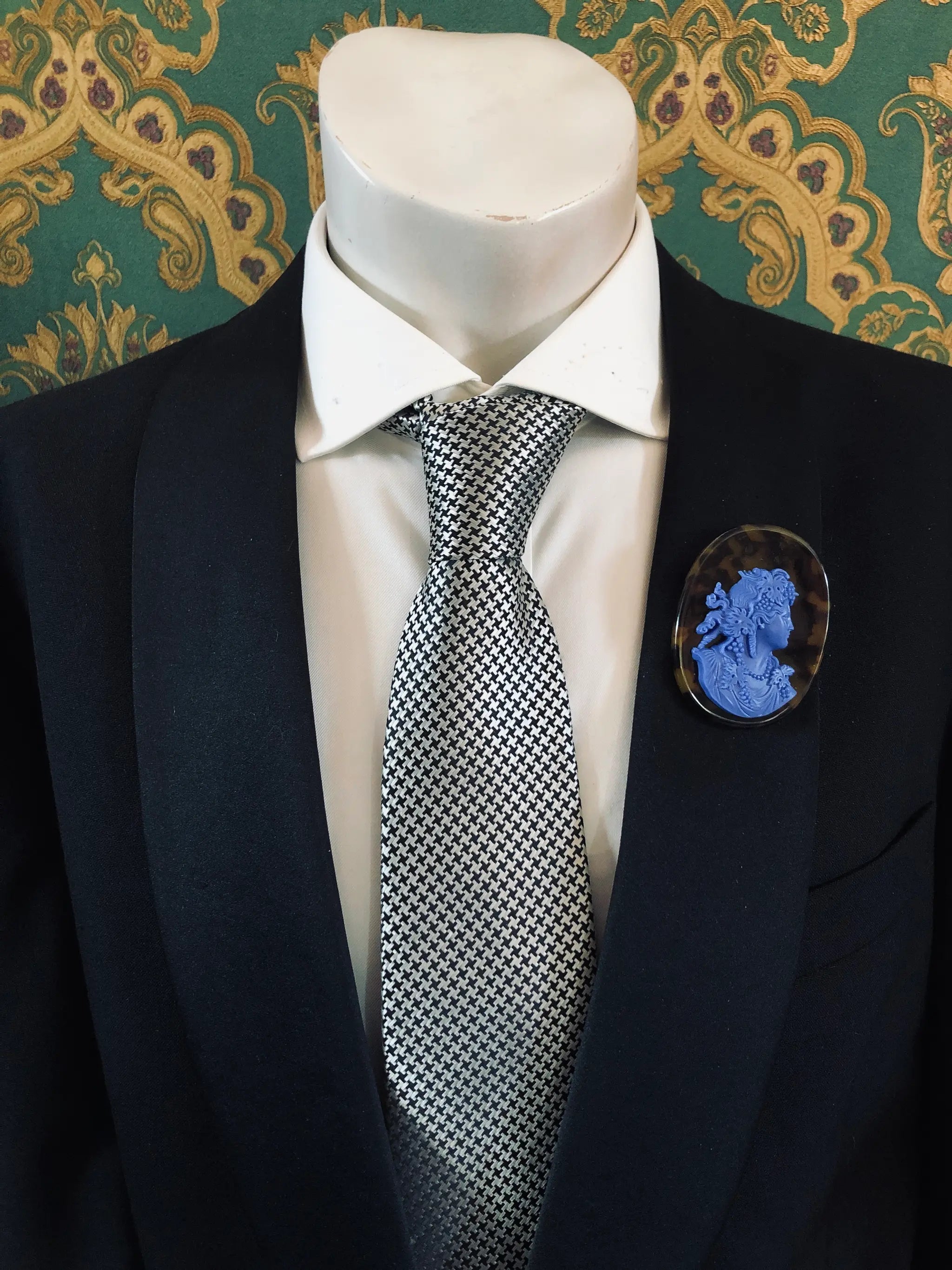 A black suit jacket on a mannequin torso, styled with a white dress shirt and a silver-and-black checkered tie. A Broochella Men's La Bella Figura bright blue and tortoiseshell cameo oval brooch is pinned to the left chest.