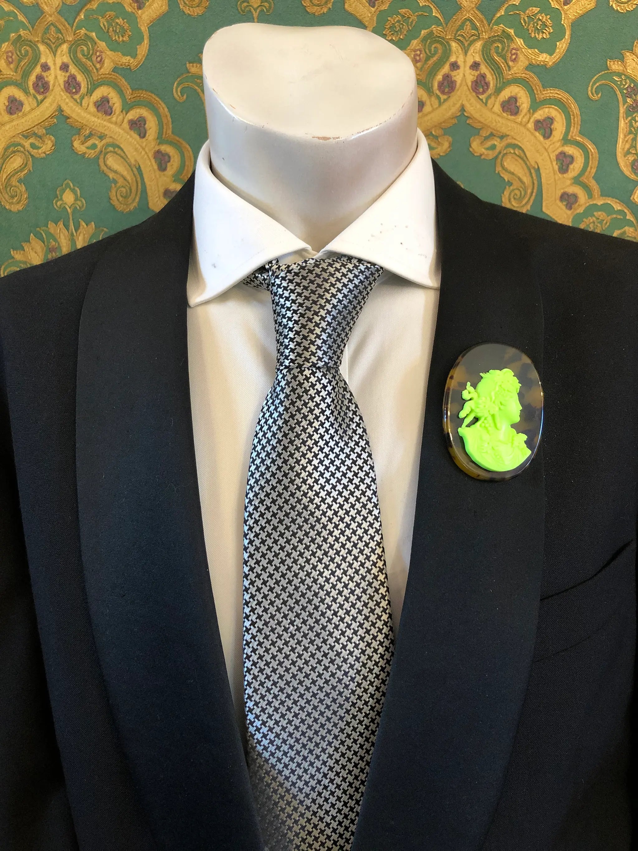 A black suit jacket on a mannequin torso, styled with a white dress shirt and a silver-and-black checkered tie. A Broochella Men's La Bella Figura bright neon green and tortoiseshell cameo oval brooch is pinned to the left chest.