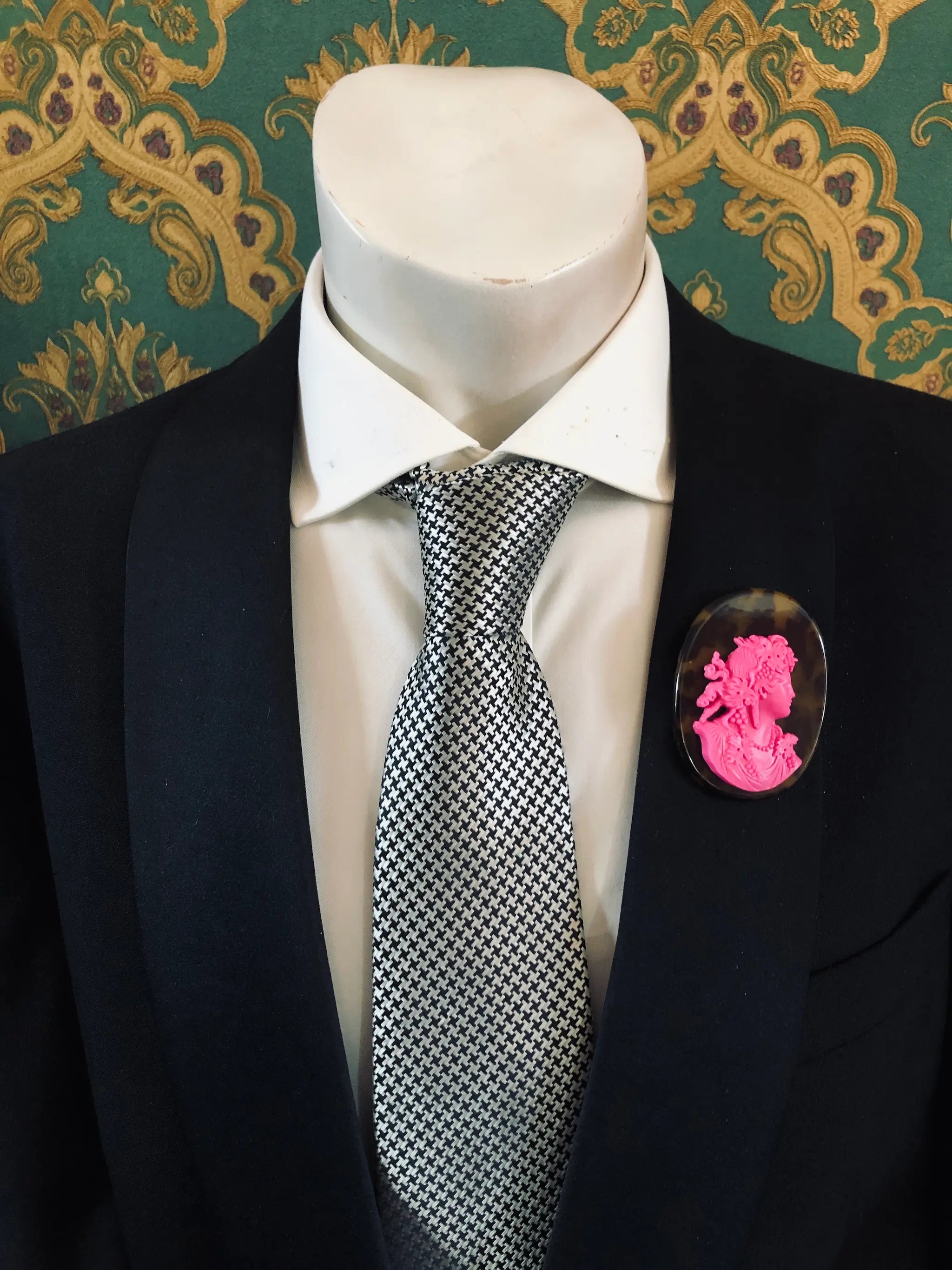 A black suit jacket on a mannequin torso, styled with a white dress shirt and a silver-and-black checkered tie. A Broochella Men's La Bella Figura bright neon green and tortoiseshell cameo oval brooch is pinned to the left chest.