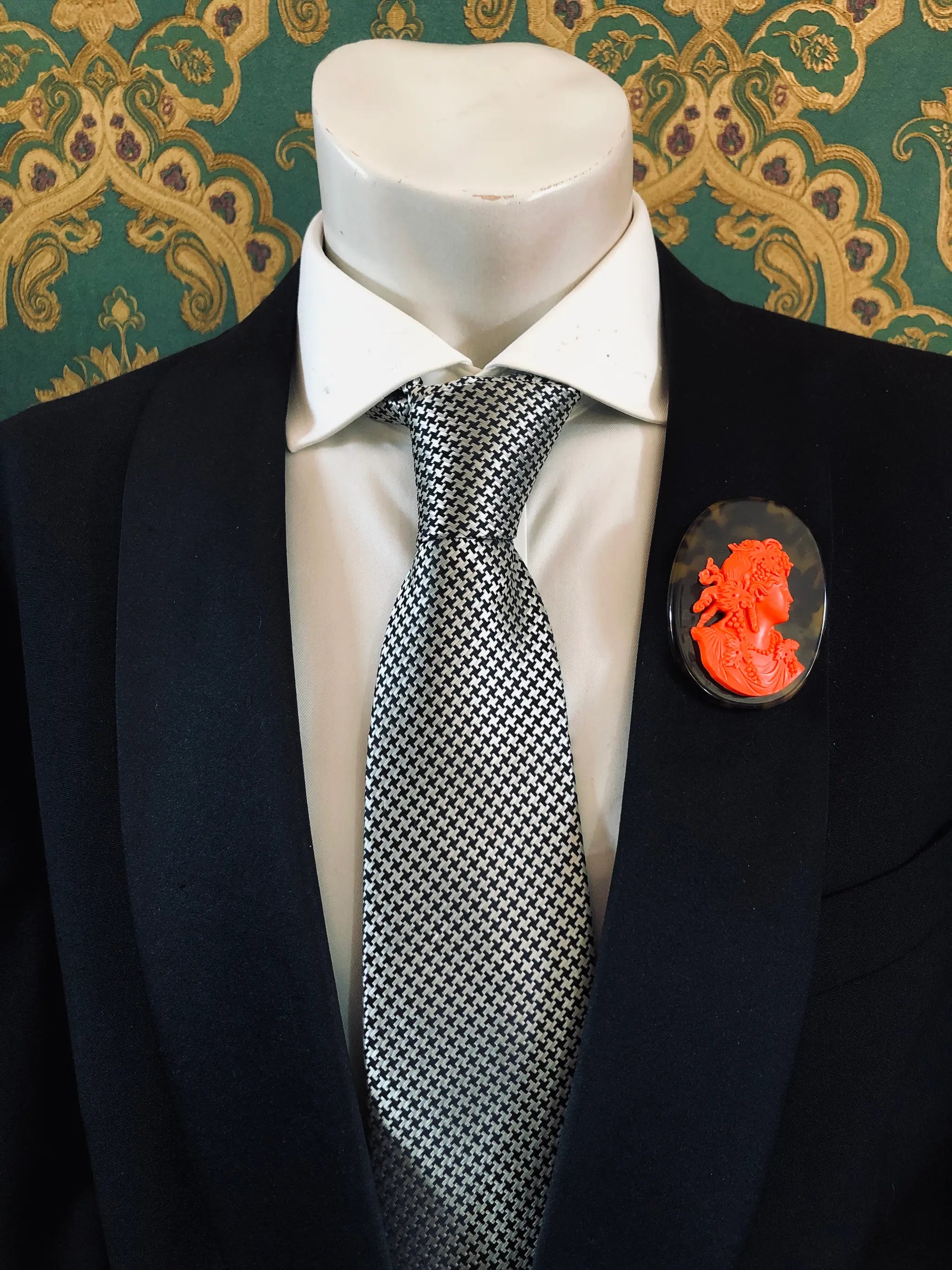 A black suit jacket on a mannequin torso, styled with a white dress shirt and a silver-and-black checkered tie. A Broochella Men's La Bella Figura bright red and tortoiseshell cameo oval brooch is pinned to the left chest.