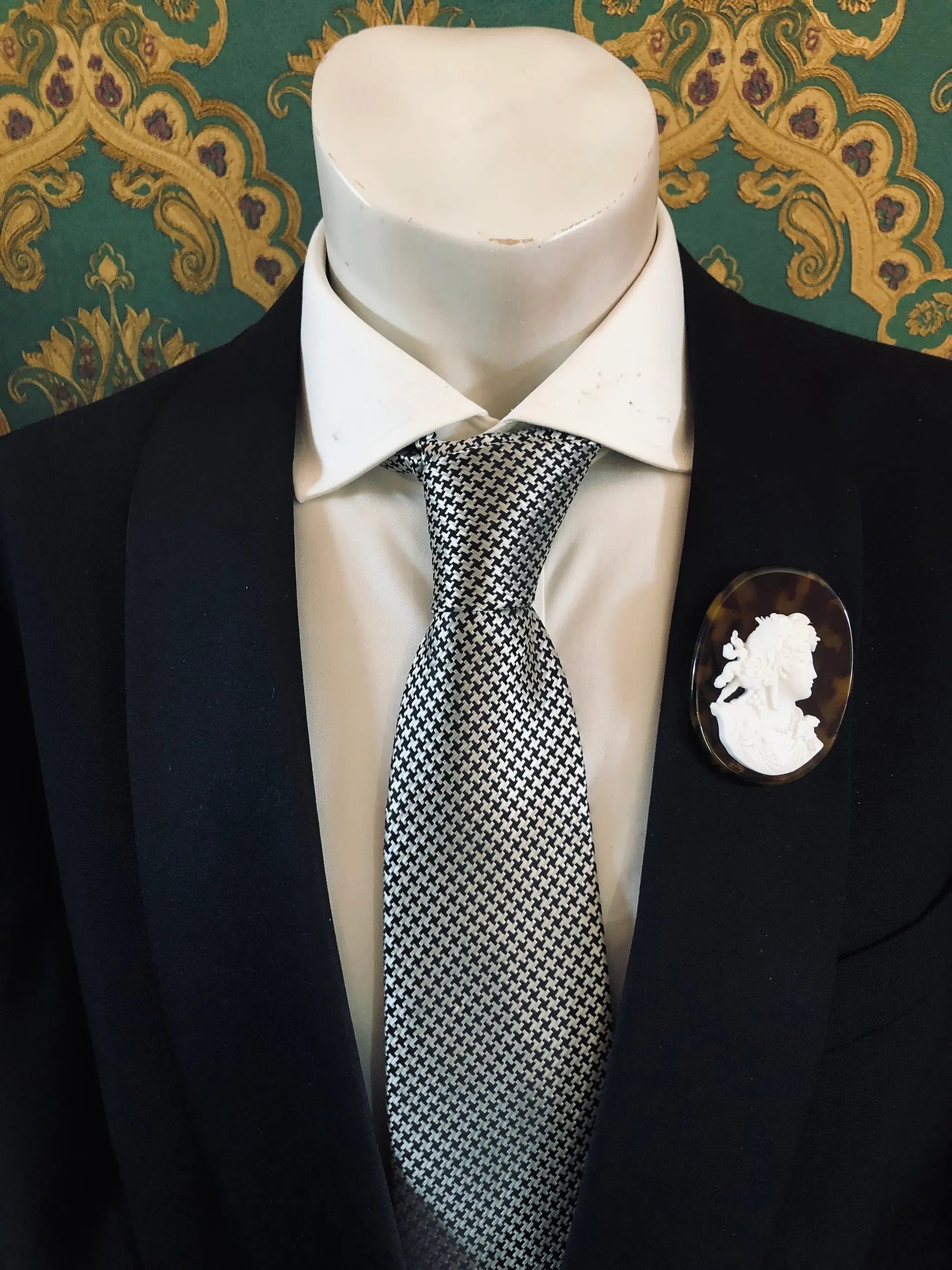 A black suit jacket on a mannequin torso, styled with a white dress shirt and a silver-and-black checkered tie. A Broochella Men's La Bella Figura white and tortoiseshell cameo oval brooch is pinned to the left chest.