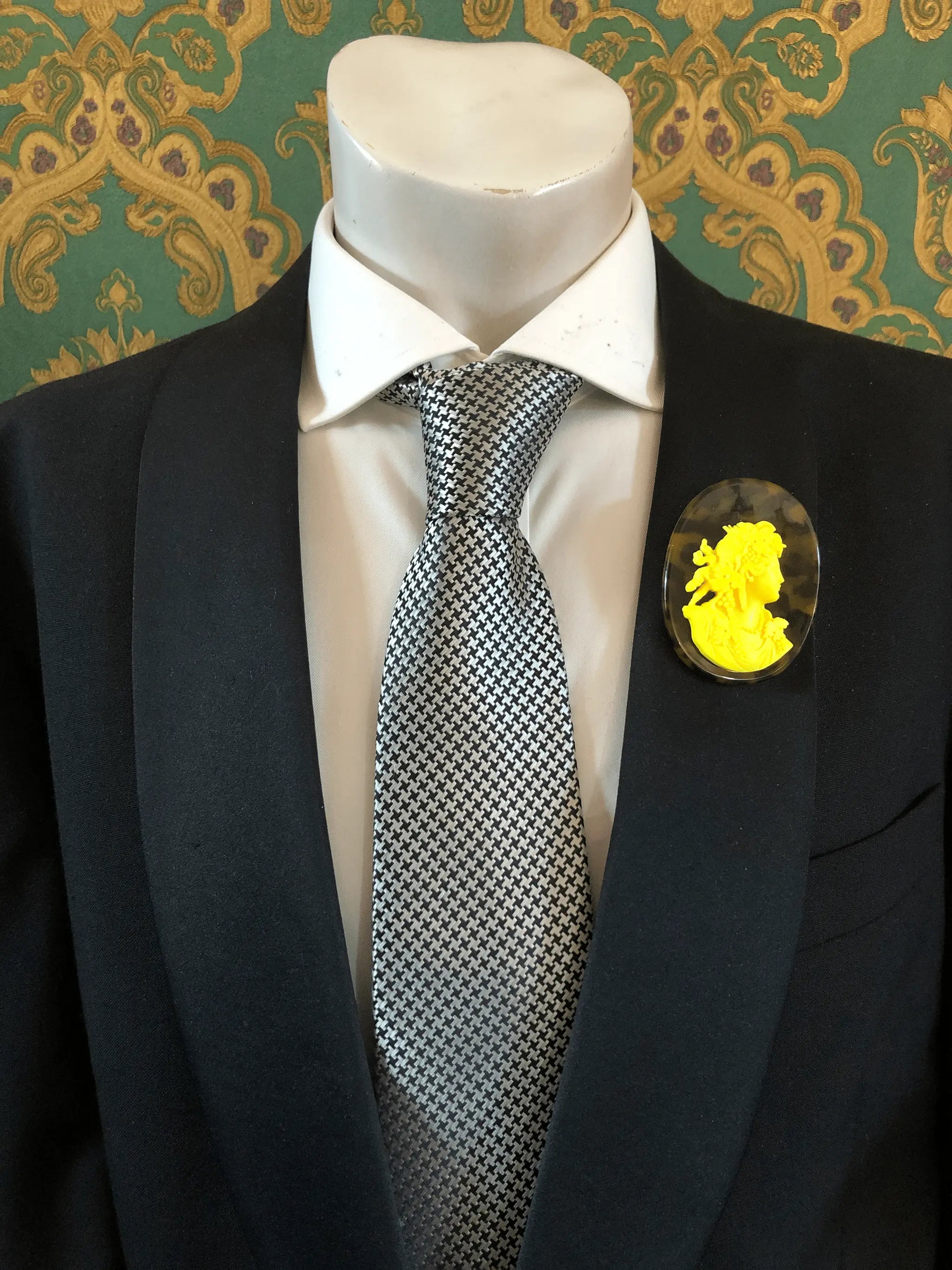 A black suit jacket on a mannequin torso, styled with a white dress shirt and a silver-and-black checkered tie. A Broochella Men's La Bella Figura bright yellow and tortoiseshell cameo oval brooch is pinned to the left chest.