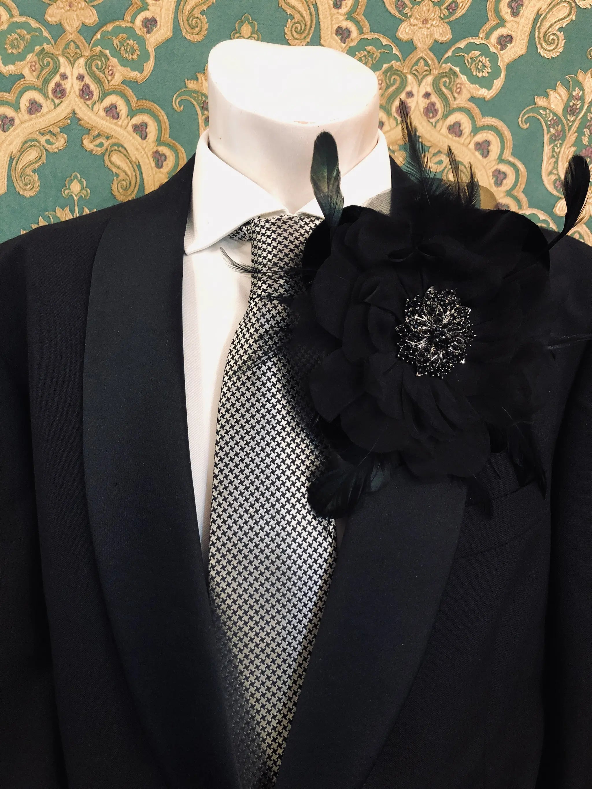 A Broochella La Contesssa black flower  brooch with feather accents and a jewelled centre. It is pinned to a black suit jacket over a white dress shirt and patterned grey tie on a mannequin. The background is green and gold damask wallpaper