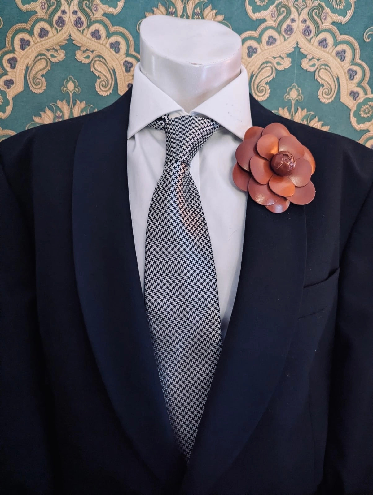 A mannequin torso wearing a black suit jacket, white dress shirt, and patterned gray tie, with a Broochella men's camelia brown leather flower brooch on the left lapel. Ornate green and gold patterned wallpaper is visible in the background.