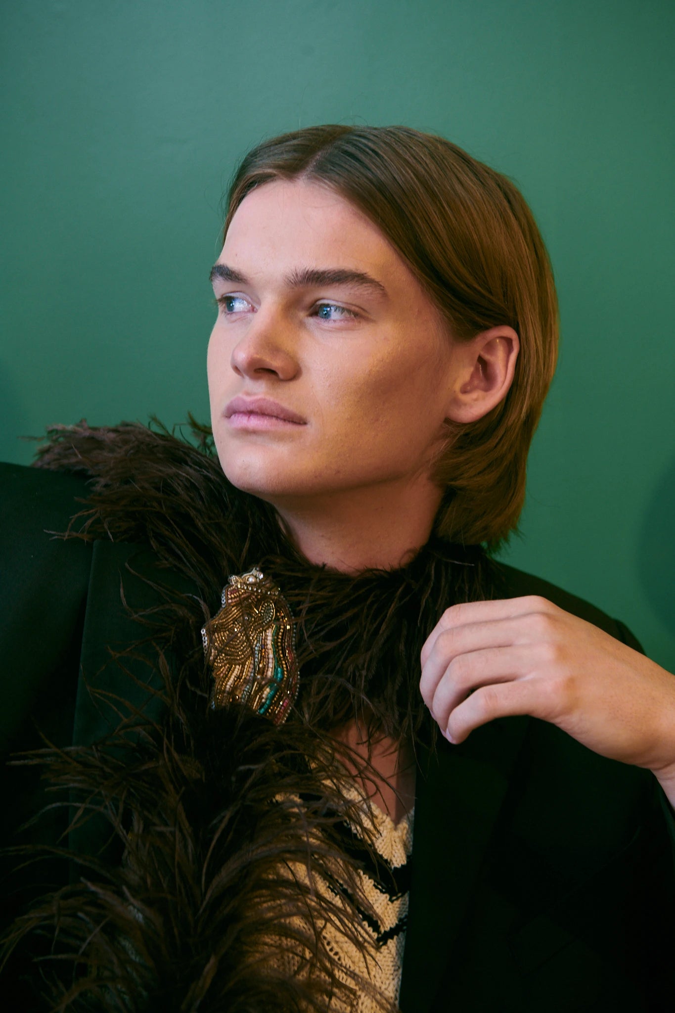 A close-up portrait of a model with light brown hair wearing a black jacket with a dark brown feather collar and a Broochella zodiac Leo large gold, decorative, hand beaded, lion brooch. It is set against a solid green background.