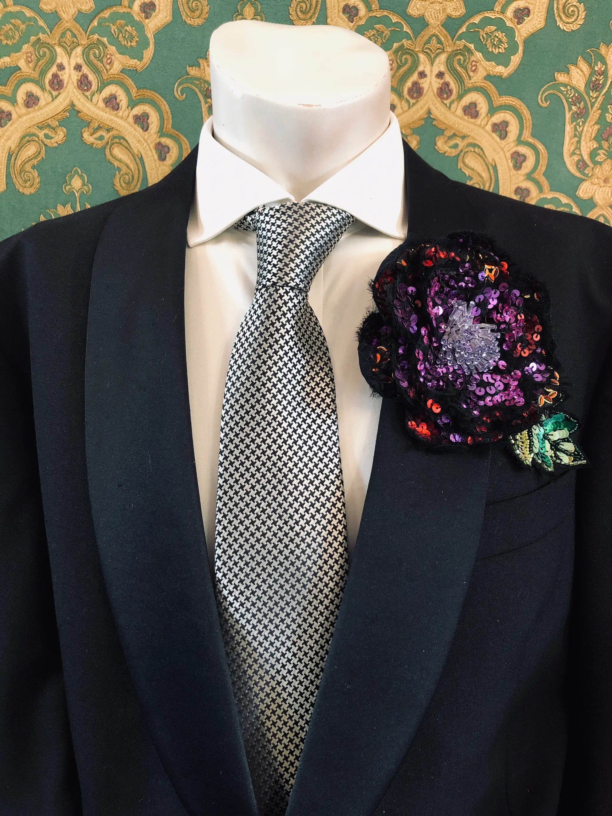 A close-up of a formal outfit on a mannequin torso, featuring a black suit jacket, white dress shirt, patterned grey and white tie, and a Broochella Men's Luminosa red, purple and orange sequinned floral hand made flower brooch. The background is green and gold damask wallpaper. 
