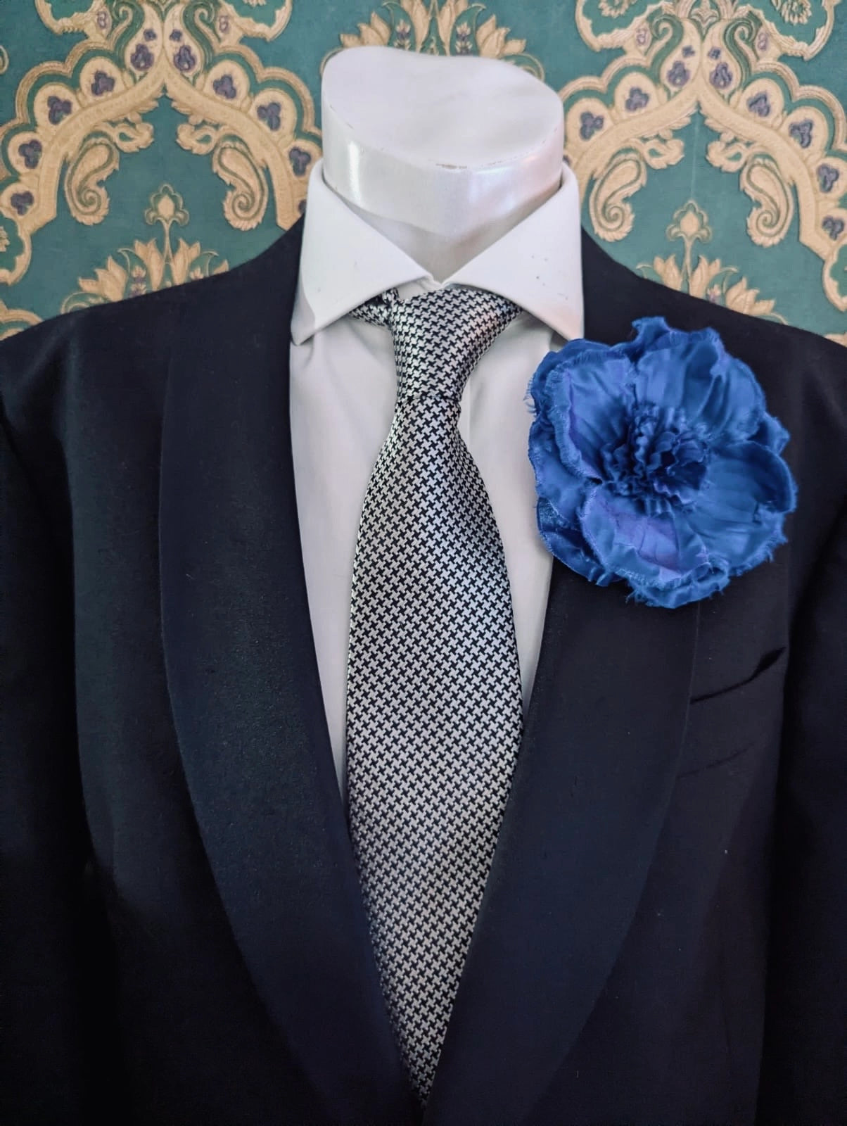 Mannequin wearing a black suit with a checkered tie and blue Broochella men's malva flower brooch against a decorative wall.