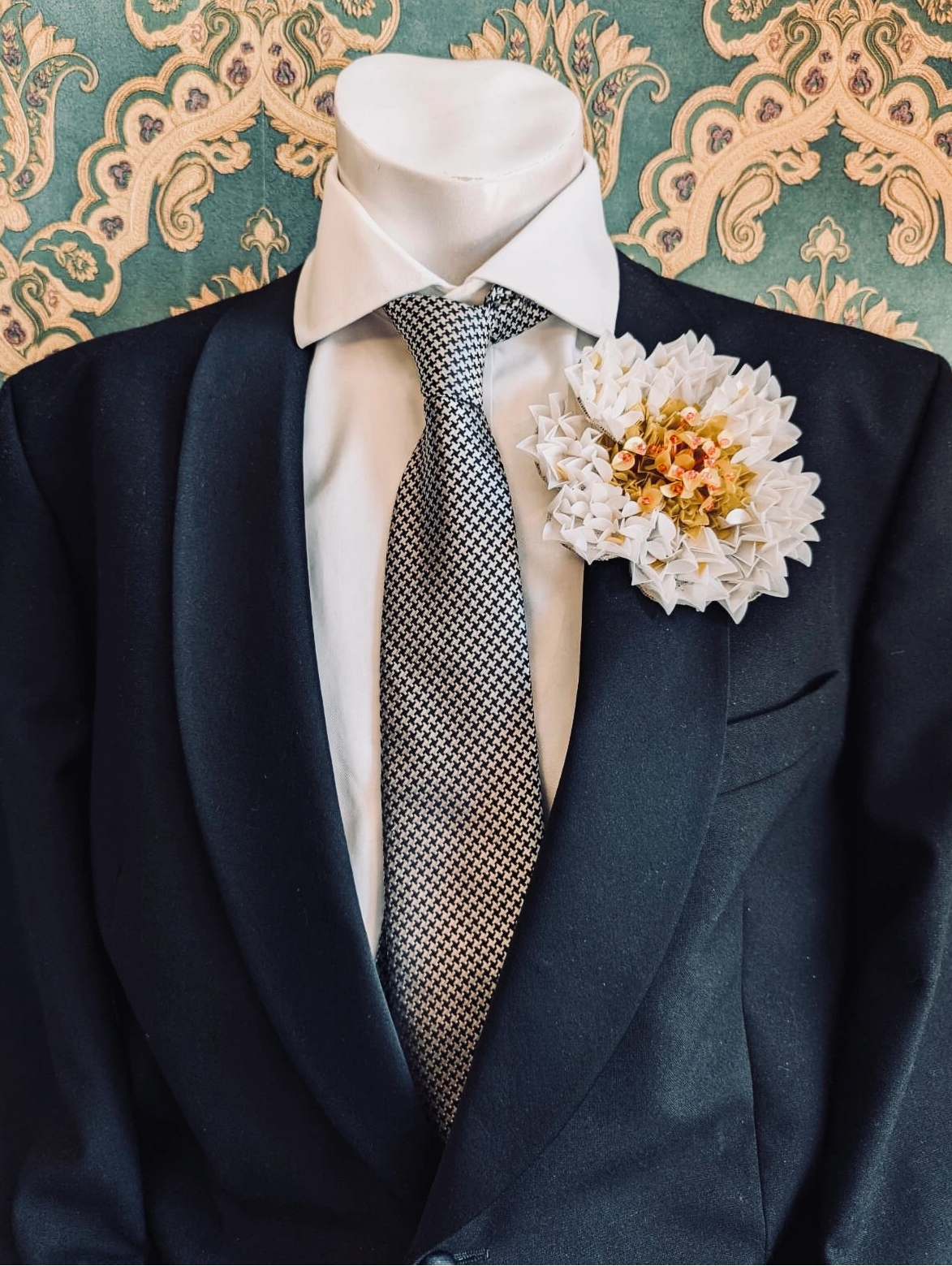 A black tuxedo jacket displayed on a white mannequin torso, styled with a white dress shirt, a grey patterned tie, and a Broochella men's handmade white narciso chrysanthemum flower brooch against a green damask patterned background.