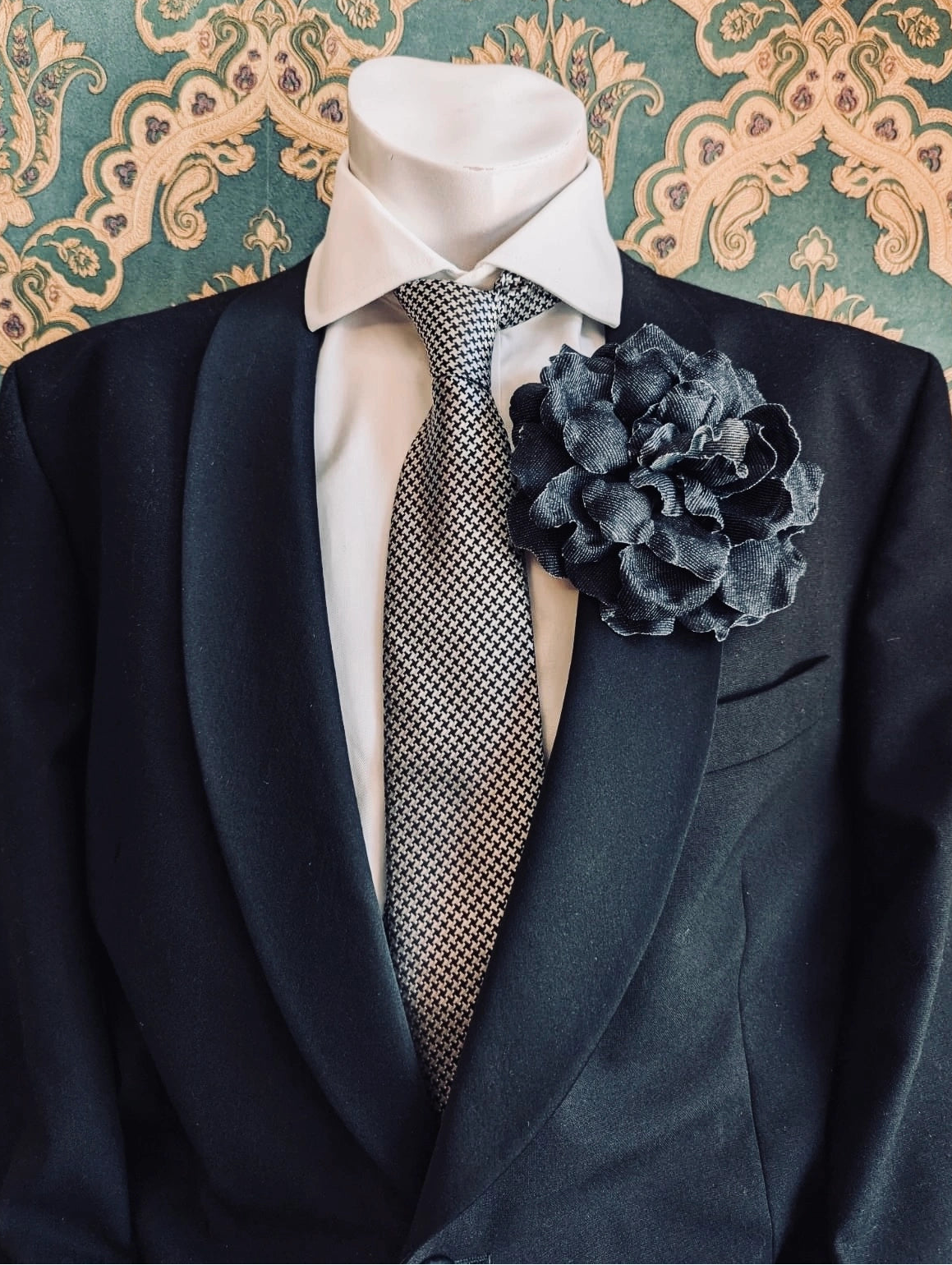 A dark navy suit jacket displayed on a white mannequin torso, styled with a white dress shirt, a grey patterned tie, and a Broochella men's navy denim handmade flower brooch, set against a teal and gold damask wallpaper background.