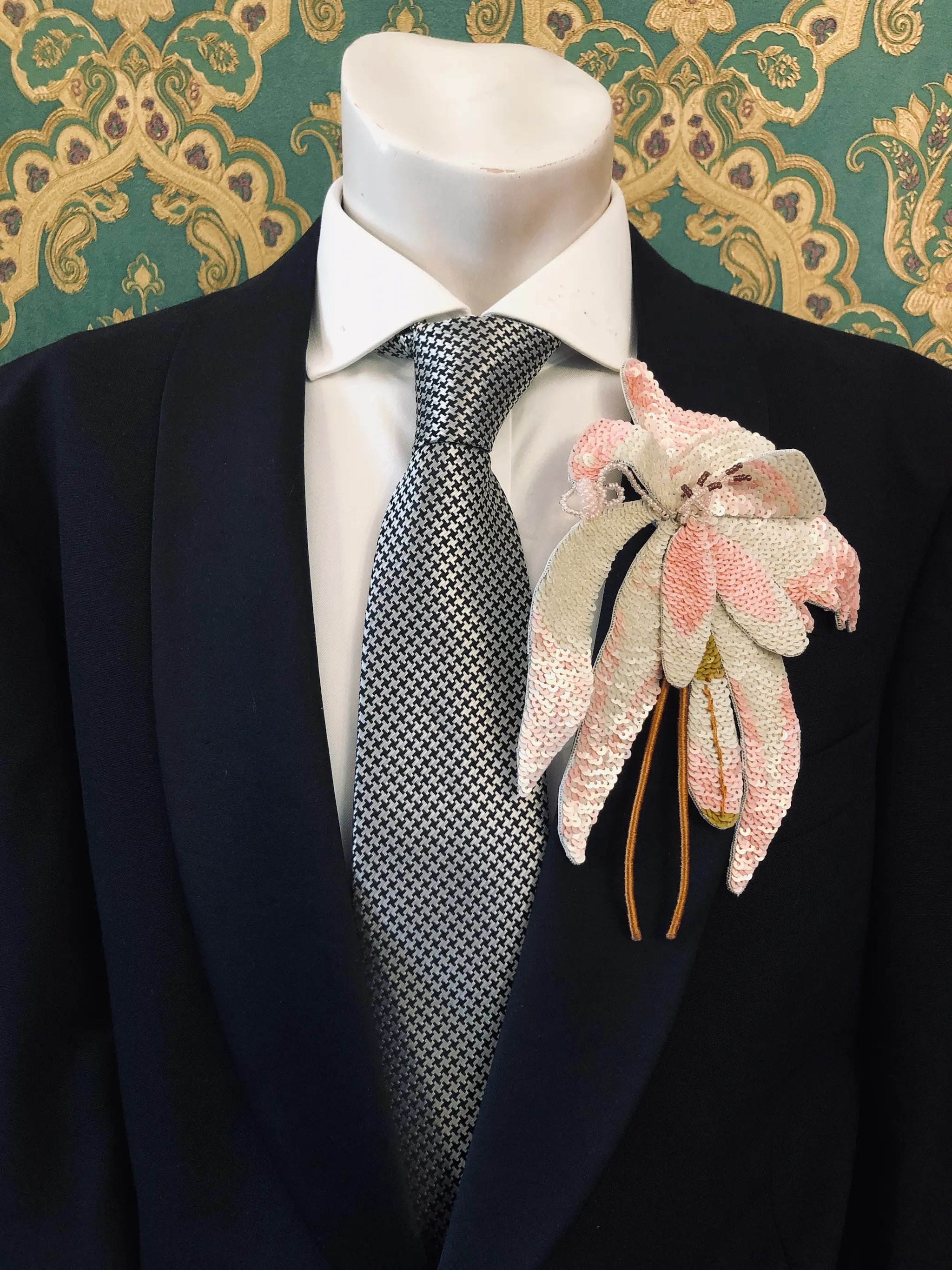 A close-up of a black suit jacket on a mannequin torso, styled with a white dress shirt, a grey patterned tie Pinned to the mannequin is the Broochella Men's Orchid flower brooch pin in pale pink, cream, and gold tones against a green damask-patterned background.