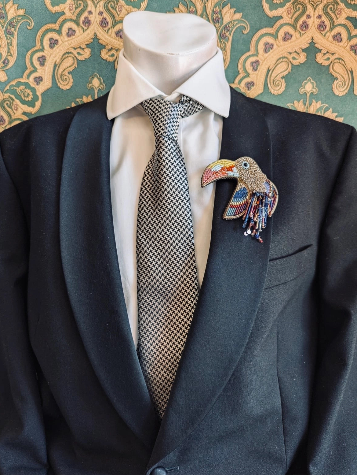 A blacksuit jacket displayed on a mannequin torso, styled with a white dress shirt, a grey patterned tie, and a colourful Broochella pappagallo parrot toucan bird lapel brooch pin against a green and gold patterned wallpaper background.