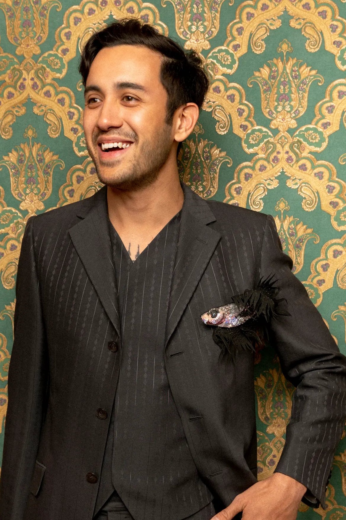 A man wearing a dark gray pinstripe three-piece suit and matching vest, standing against a green and gold damask patterned backdrop. He is wearing the Broochella men's siamese fish brooch. He is smiling and looking to the side, with one hand in his pocket and the other holding the lapel of his jacket.