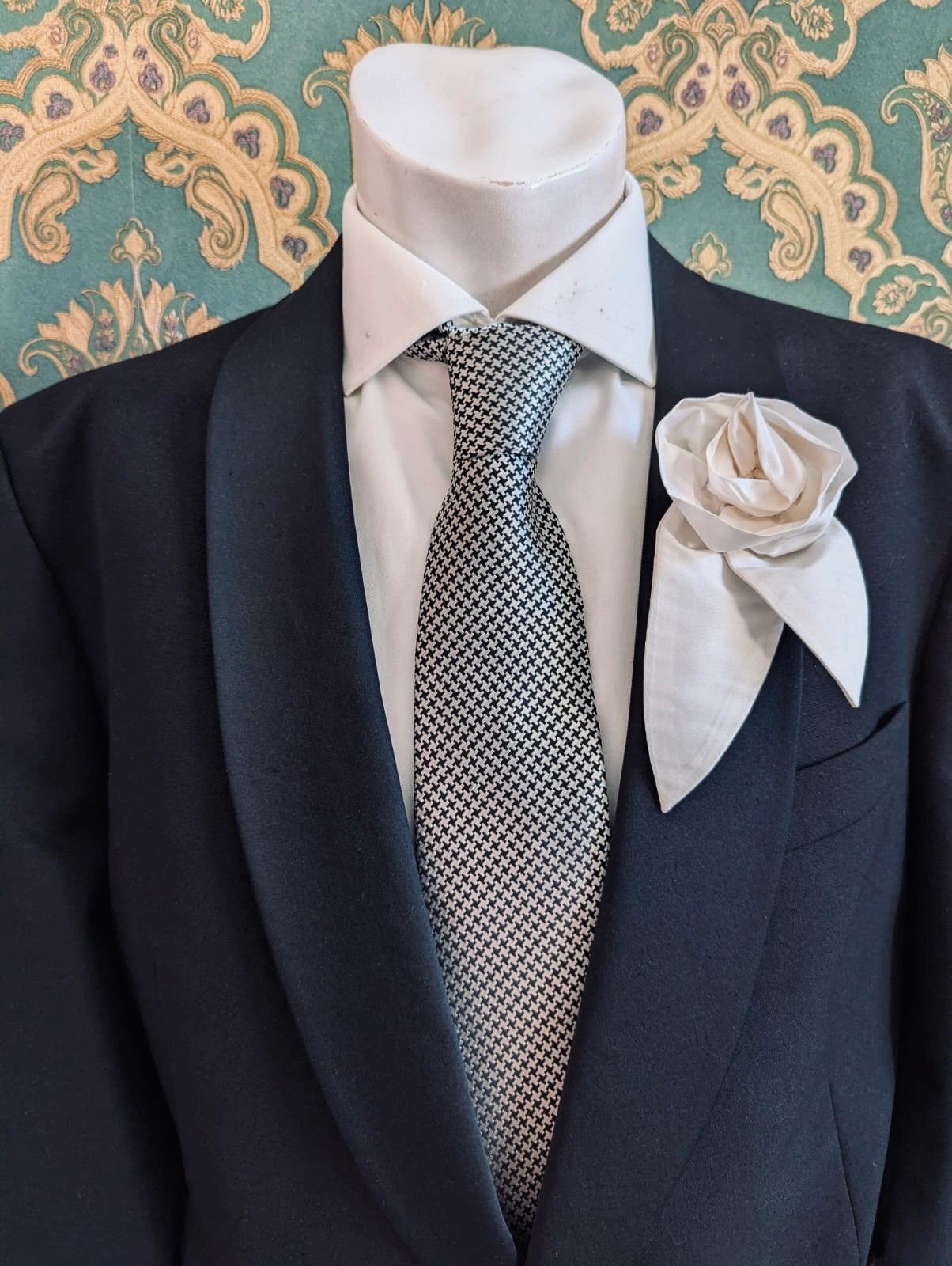 A Broochella Men's Petalo ivory, small, handmade silk  rose-shaped flower brooch with petals. It is displayed on a men's mannequin that is dress in a black suit, with a silver and black houndstooth tie. It is against a dark teal and gold damask patterned wallpaper background