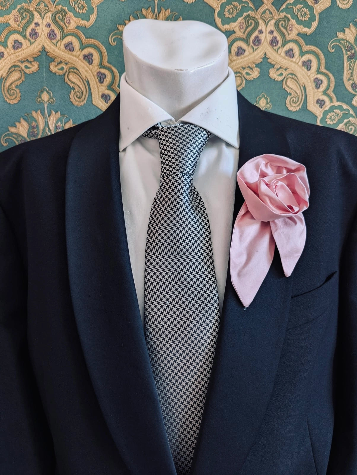 A Broochella Men's Petalo pink, small, handmade silk  rose-shaped flower brooch with petals. It is displayed on a men's mannequin that is dress in a black suit, with a silver and black houndstooth tie. It is against a dark teal and gold damask patterned wallpaper background