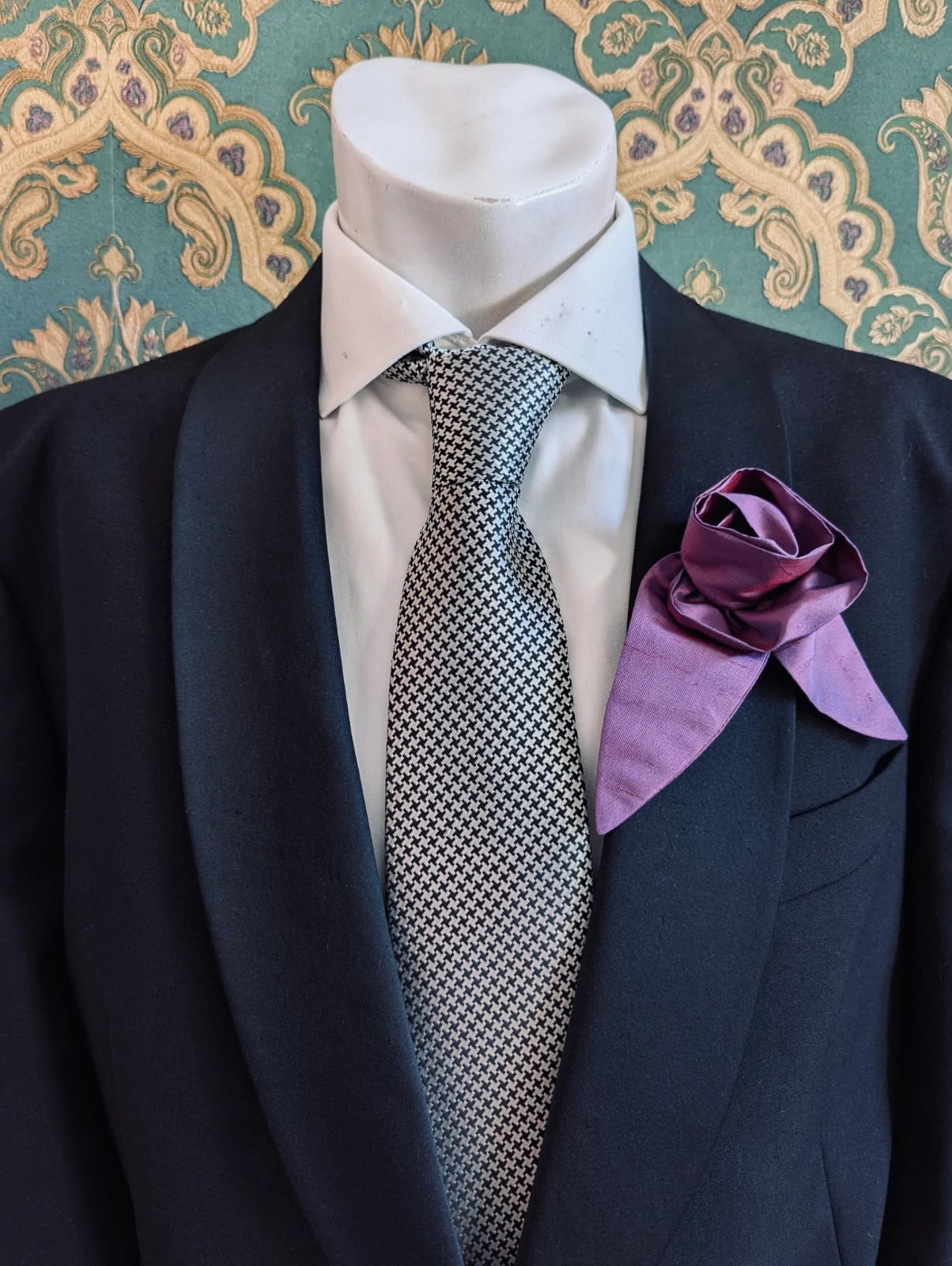 A Broochella Men's Petalo purple, small, handmade silk  rose-shaped flower brooch with petals. It is displayed on a men's mannequin that is dress in a black suit, with a silver and black houndstooth tie. It is against a dark teal and gold damask patterned wallpaper background