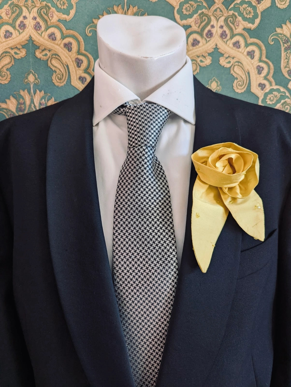 A Broochella Men's Petalo yellow, small, handmade silk  rose-shaped flower brooch with petals. It is displayed on a men's mannequin that is dress in a black suit, with a silver and black houndstooth tie. It is against a dark teal and gold damask patterned wallpaper background
