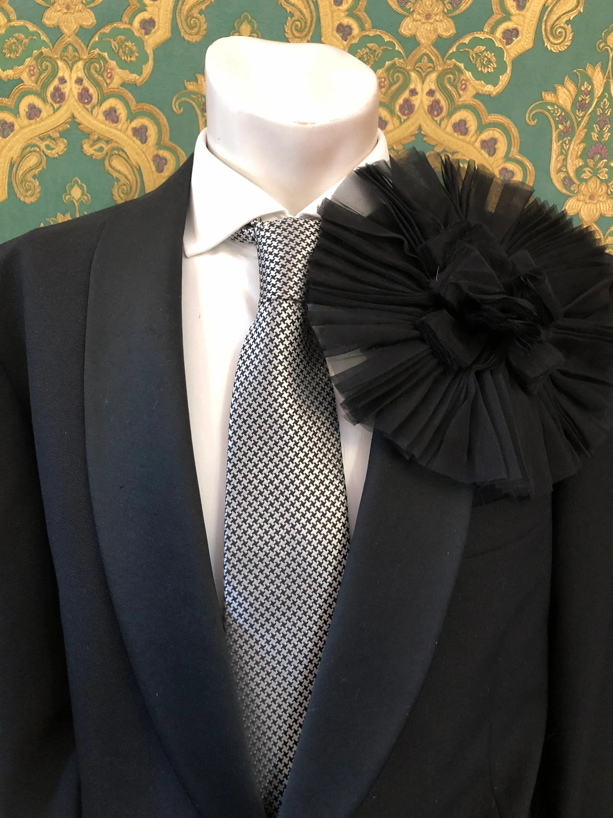 A formal outfit displayed on a mannequin torso, featuring a black suit jacket, white dress shirt, patterned grey and white tie, and a large Broochella Men's Pieghettato black flower brooch on the lapel. The background is ornate green and gold damask wallpaper.