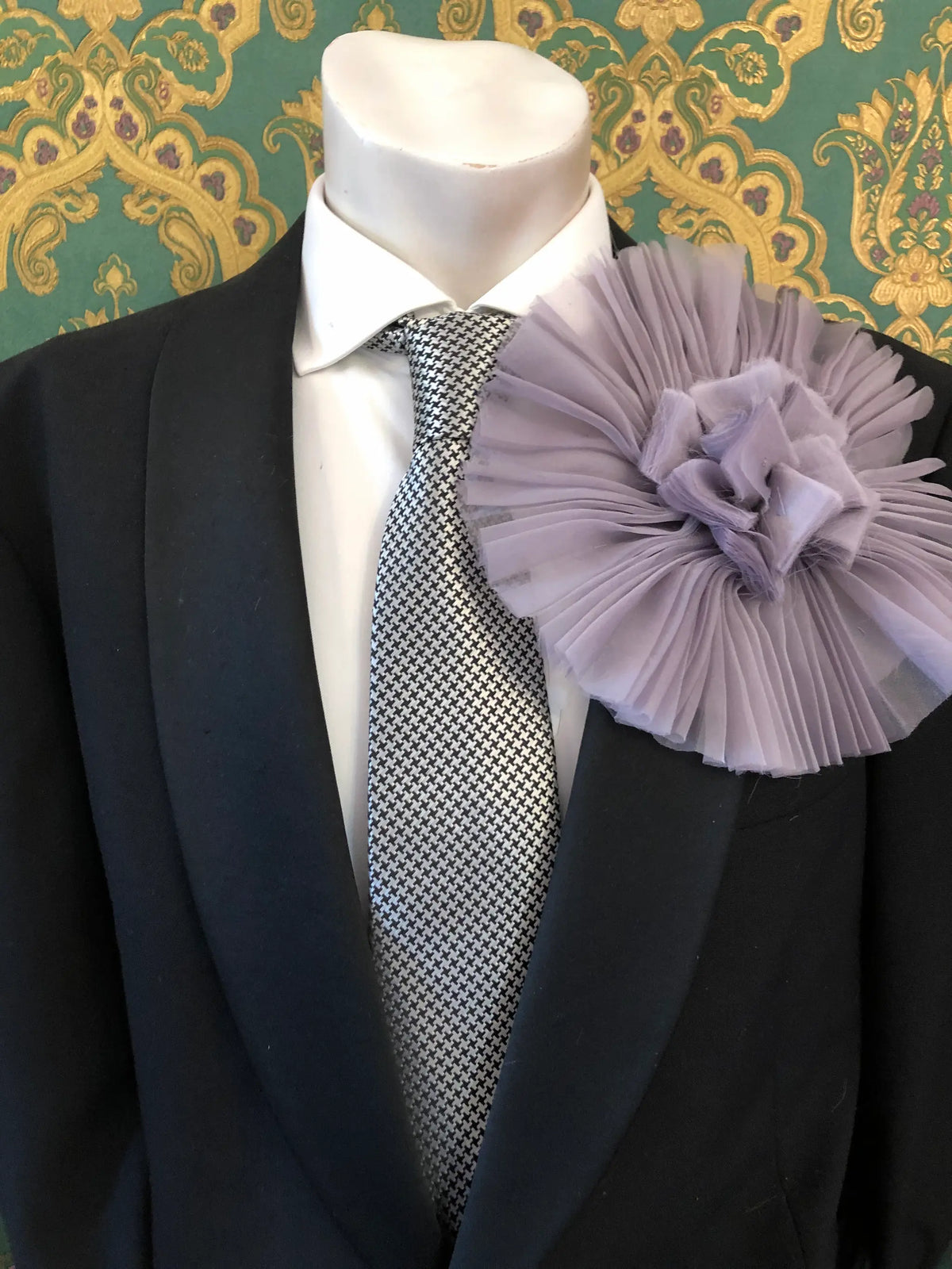 A formal outfit displayed on a mannequin torso, featuring a black suit jacket, white dress shirt, patterned grey and white tie, and a large Broochella Men's Pieghettato lavender flower brooch on the lapel. The background is ornate green and gold damask wallpaper.
