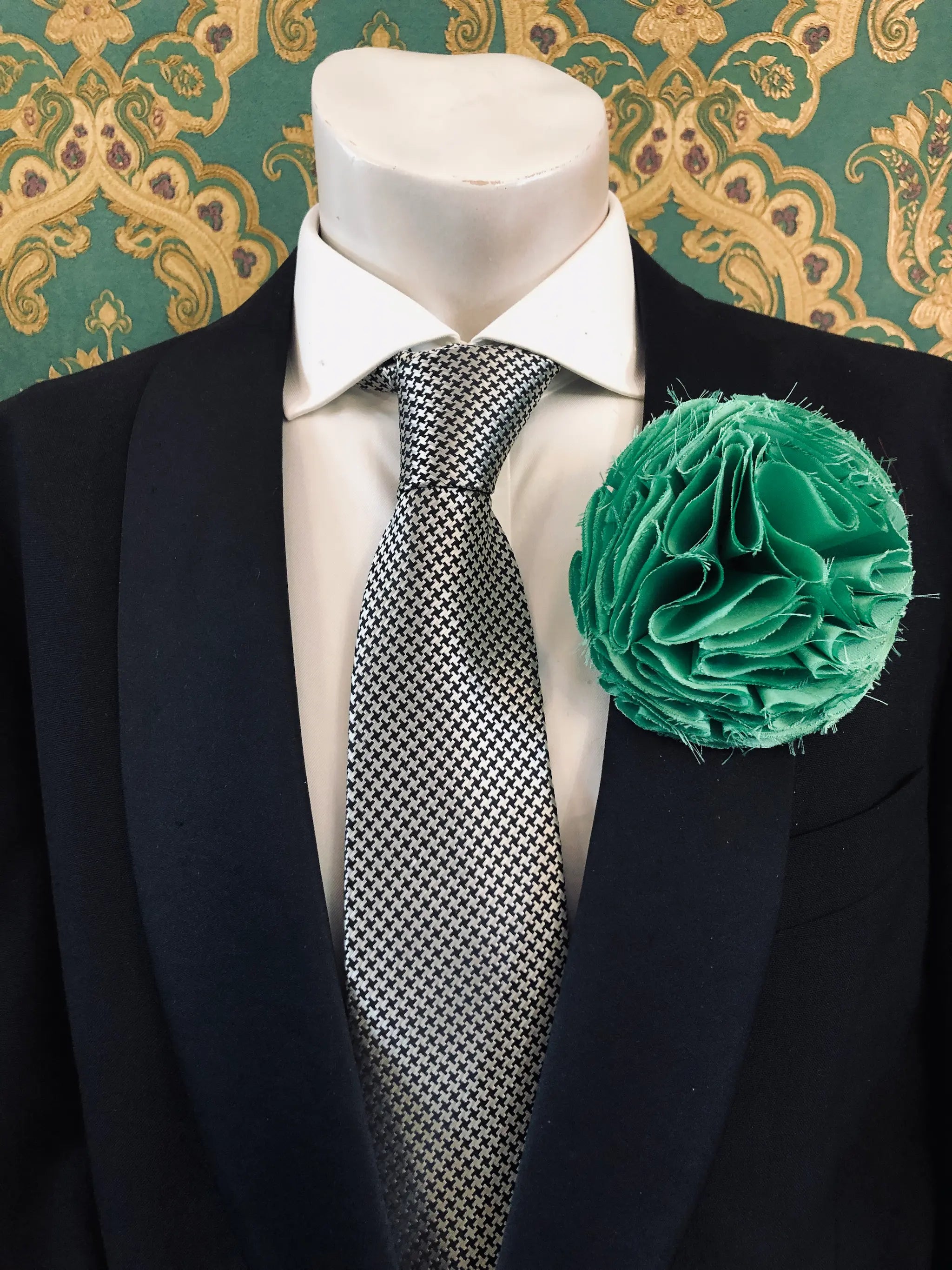 A men's formal wear ensemble displayed on a white mannequin torso, featuring a black suit jacket, a white dress shirt, a black and white checkered pattern tie, and a vibrant Broochella Men's Pom Pon green textured fabric flower brooch.