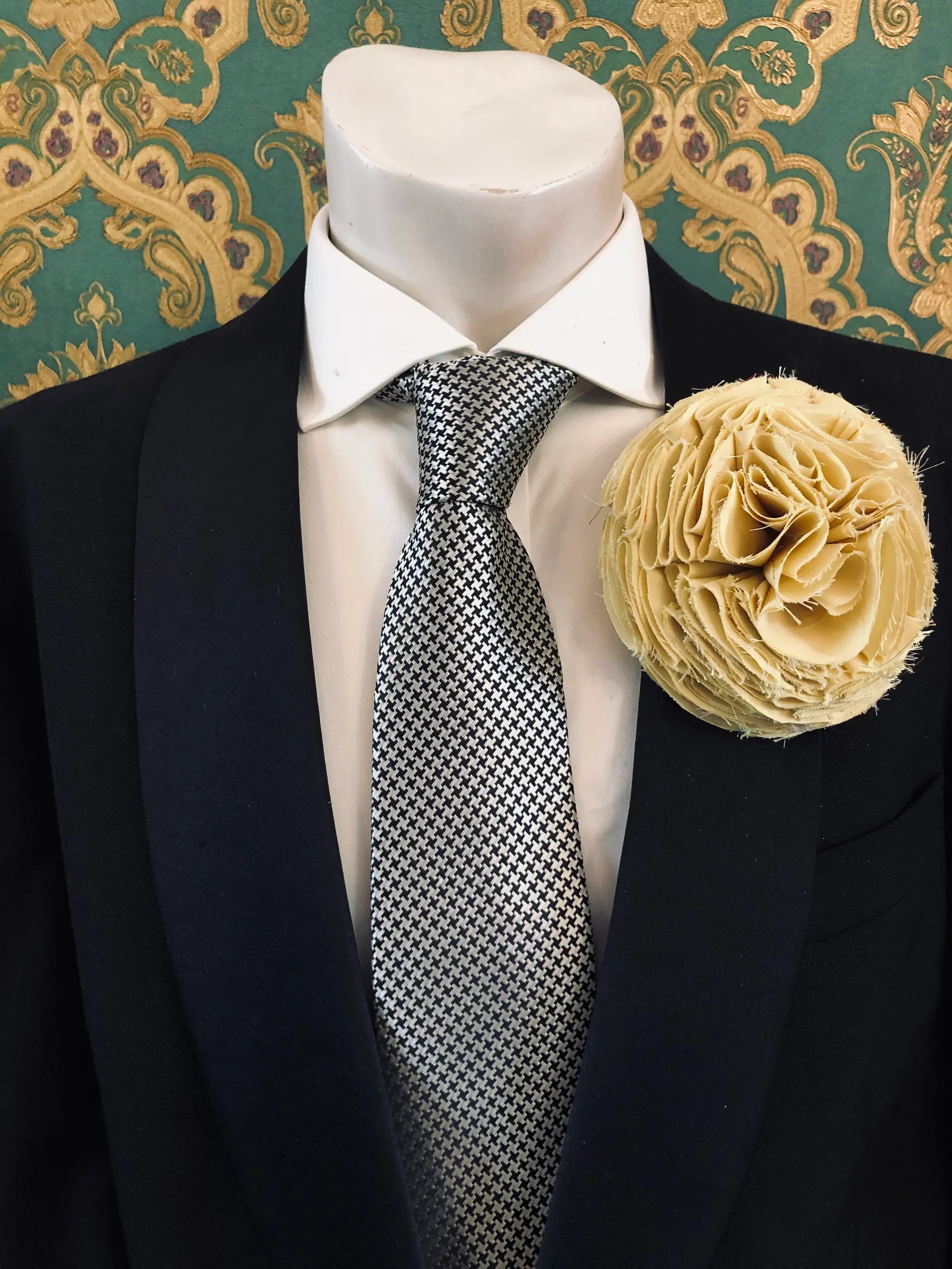 A men's formal wear ensemble displayed on a white mannequin torso, featuring a black suit jacket, a white dress shirt, a black and white checkered pattern tie, and a vibrant Broochella Men's Pom Pon yellow textured fabric flower brooch.