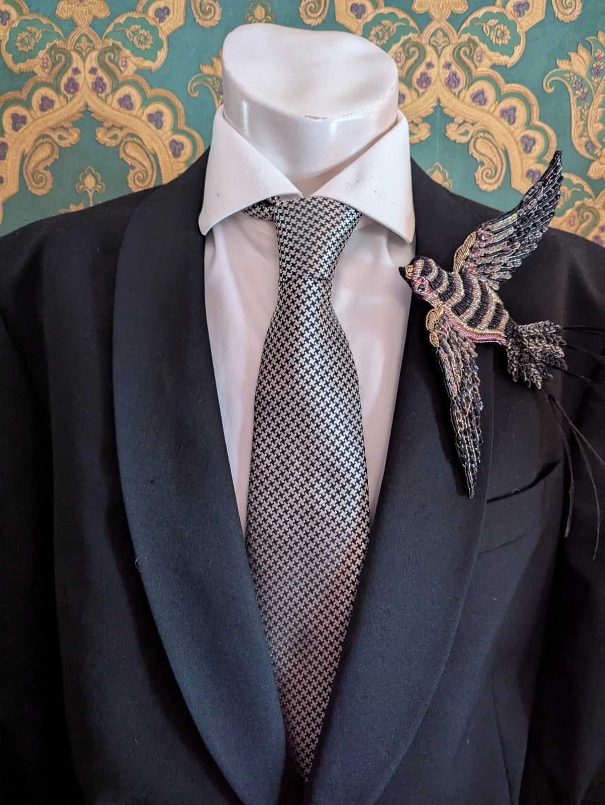 Mannequin wearing a black suit with a white shirt, patterned tie, and Broochella hand beaded designer rondine swallow bird decorative brooch against a green and gold damask wallpapered wall.