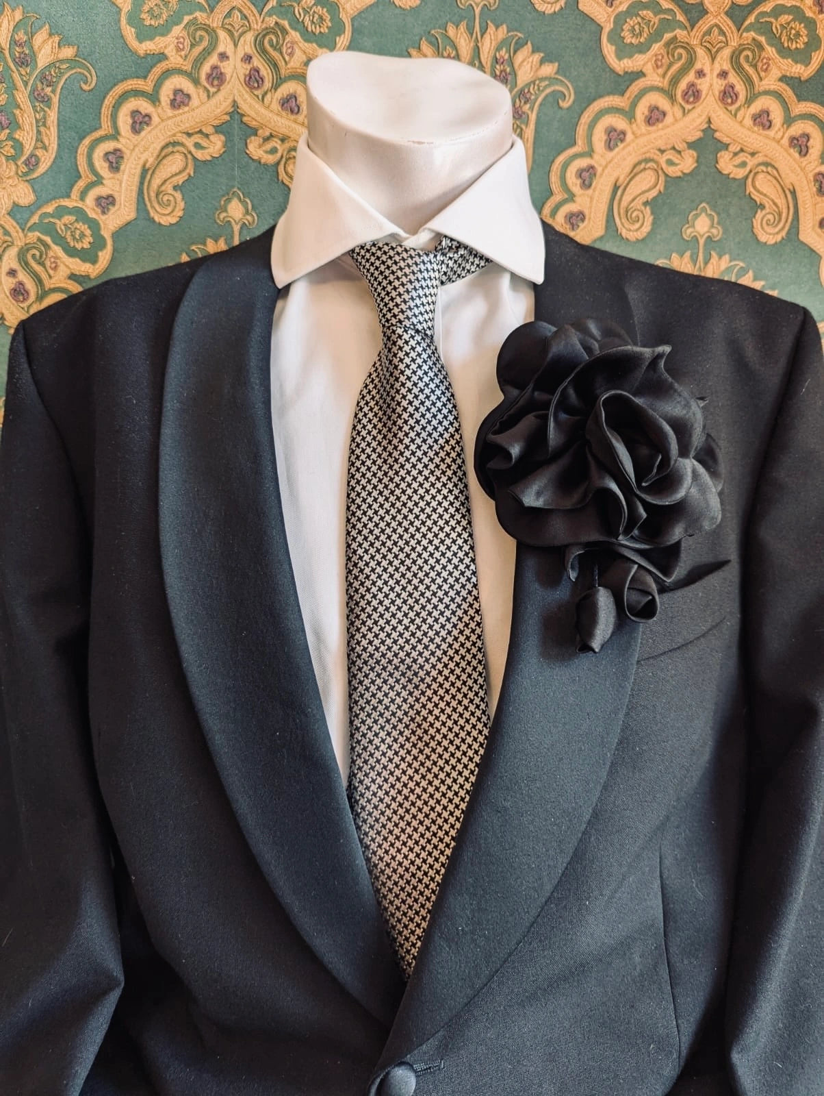 Broochella men's rosetta handmade, designer, satin flower brooch in black. Pinned on a men's mannequin wearing a black suit and a houndstooth tie. The background is green and gold damask wallpaper. 