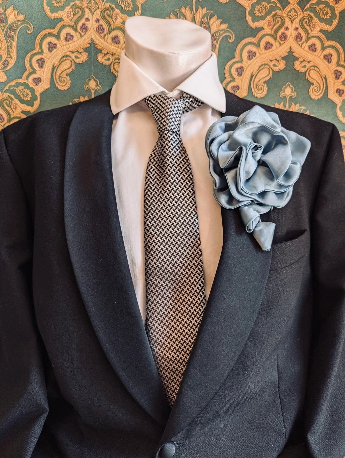 Broochella men's rosetta handmade, designer, satin flower brooch in blue. Pinned on a men's mannequin wearing a black suit and a houndstooth tie. The background is green and gold damask wallpaper. 