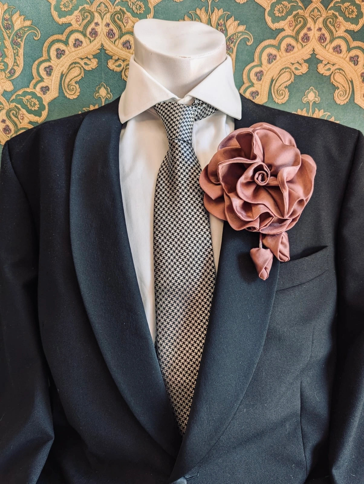 Broochella men's rosetta handmade, designer, satin flower brooch in blush pink. Pinned on a men's mannequin wearing a black suit and a houndstooth tie. The background is green and gold damask wallpaper. 