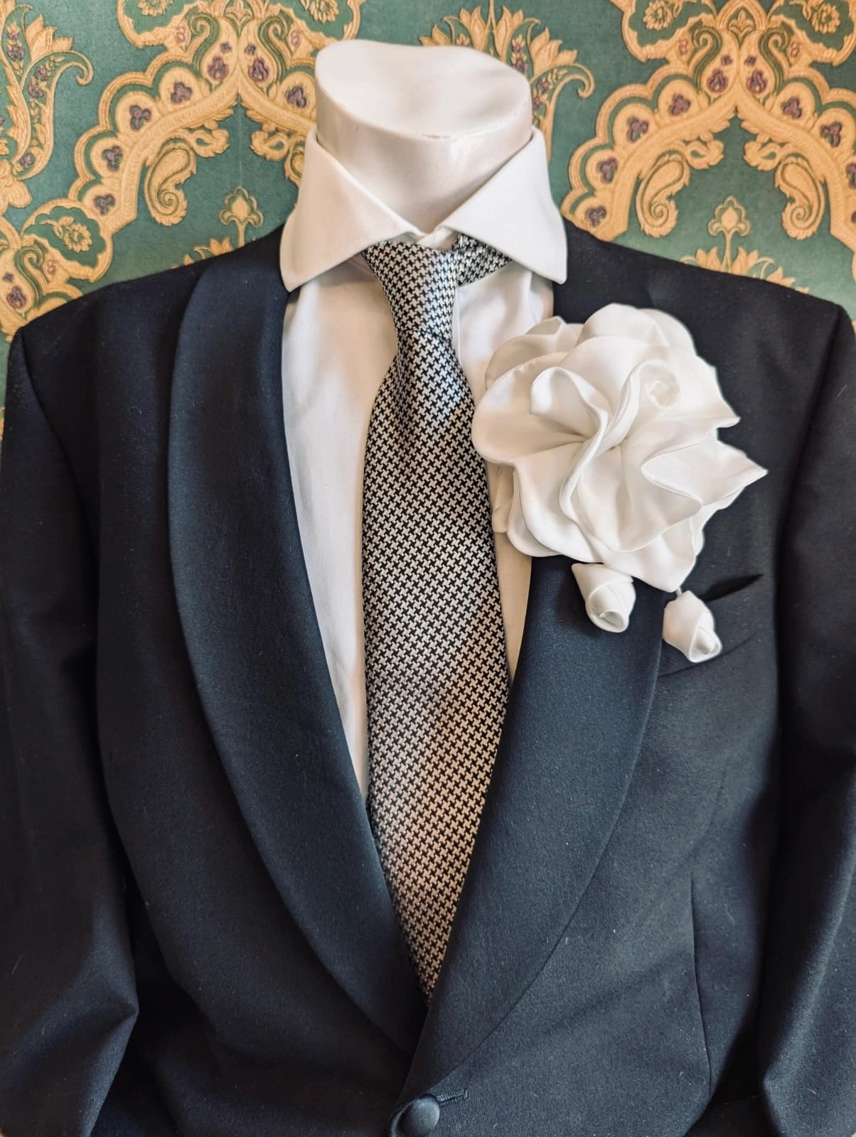 Broochella men's rosetta handmade, designer, satin flower brooch in white. Pinned on a men's mannequin wearing a black suit and a houndstooth tie. The background is green and gold damask wallpaper. 