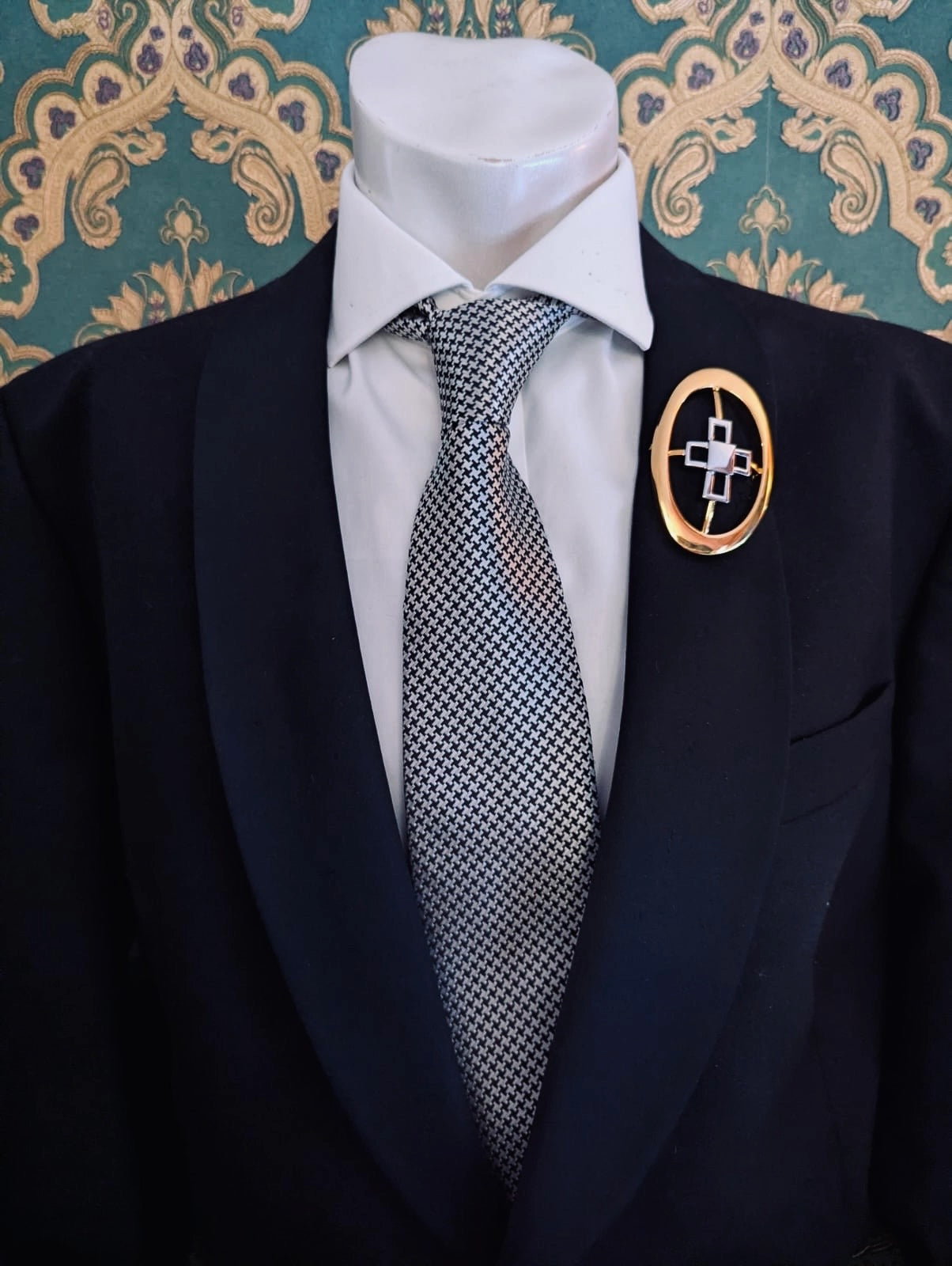 Broochella men's brooches with meaning knowledge gold metal and silver brooch. Pinned to a men's mannequin wearing a black suit and houndstooth tie. The background is green and gold damask wallpaper.
